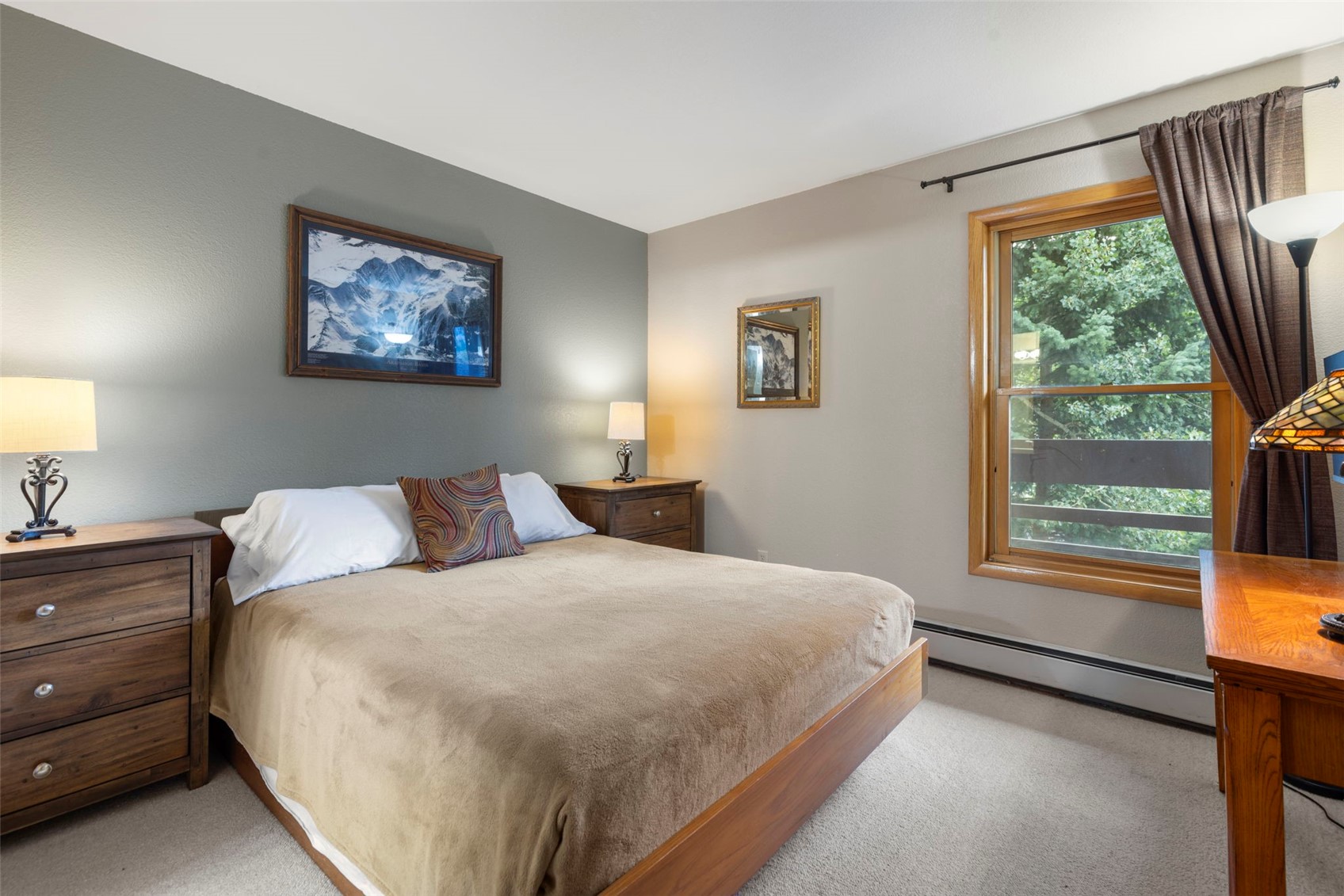 This cozy bedroom features a central bed with a tan blanket and a patterned accent pillow, flanked by two wooden nightstands topped with matching lamps. A framed mountain landscape print hangs above the bed, while a window with brown curtains provides a view of the trees outside. The room is finished with neutral wall tones and light-colored carpeting, creating a warm and inviting atmosphere.