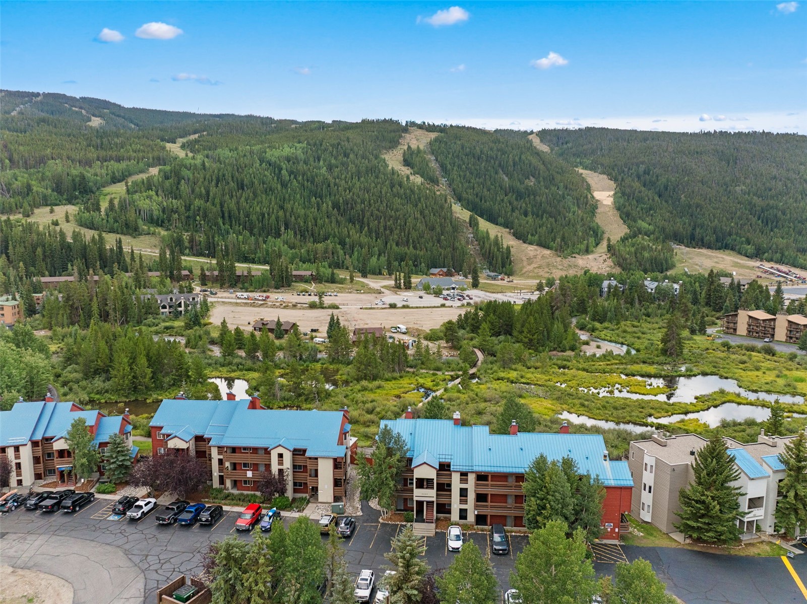This high-angle aerial shot captures a multi-building residential complex with distinctive blue metal roofs nestled in a mountainous landscape. The property features paved parking areas in the foreground, transitioning into a lush, marshy green space with small ponds, leading toward a vast, undeveloped valley and a forested ski mountain in the distance. The scene conveys a serene, recreational atmosphere typical of a mountain resort community.
