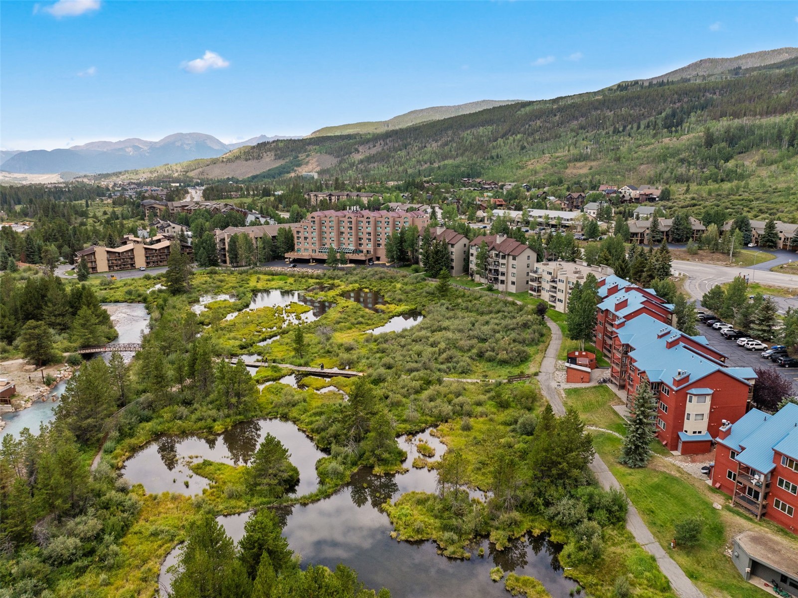 This high-angle aerial view captures a scenic mountain resort community nestled in a lush, green valley. The landscape features a prominent wetland area with winding boardwalks and small ponds, surrounded by multi-story residential buildings with blue and brown roofs. In the background, rolling hills and mountains under a clear blue sky provide a picturesque backdrop for this tranquil outdoor setting.