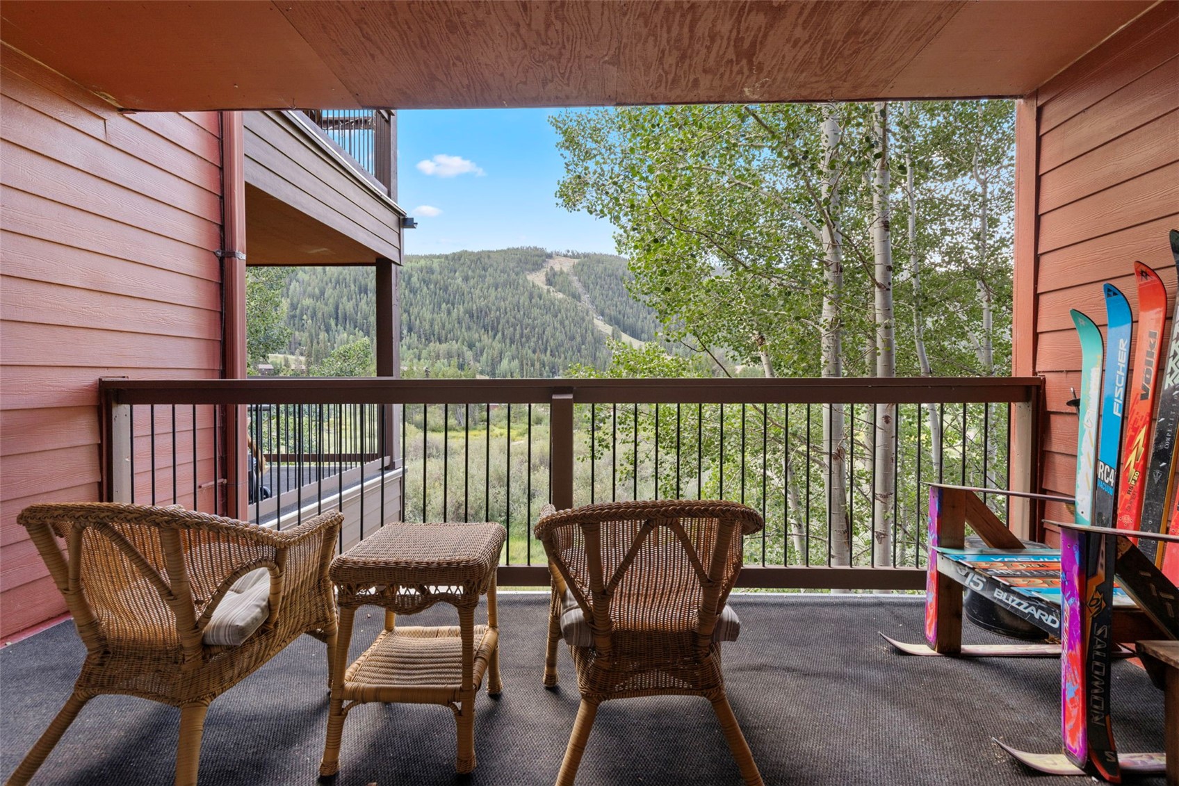 This elevated balcony offers a serene outdoor living space furnished with two wicker chairs and a matching side table. The deck features a dark railing that frames a picturesque view of a lush, tree-lined mountain landscape. Several pairs of skis are neatly stored against the side wall, highlighting the property's proximity to winter sports.