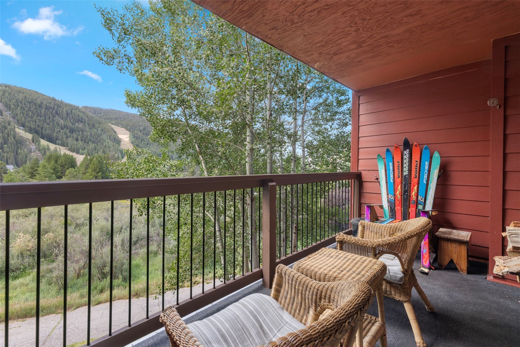 This elevated balcony offers a scenic view of a lush, tree-lined mountain landscape and a ski slope in the distance. The space is furnished with two wicker chairs and a small matching table, creating a cozy spot for outdoor relaxation. A collection of colorful skis is propped against the red-painted wooden wall, adding a touch of local character to this mountain-style retreat.