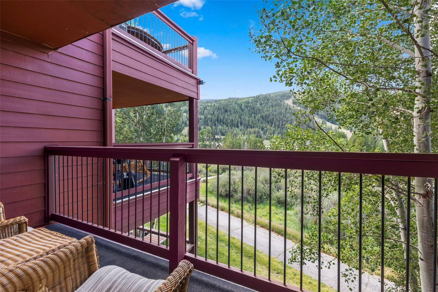 This elevated balcony features a dark red wooden exterior wall and a matching railing, providing a cozy outdoor space with wicker seating. The perspective looks out over a lush, green landscape with a paved path winding through trees toward a distant, forested mountain slope. The scene captures a serene, natural setting, perfect for enjoying the mountain views.
