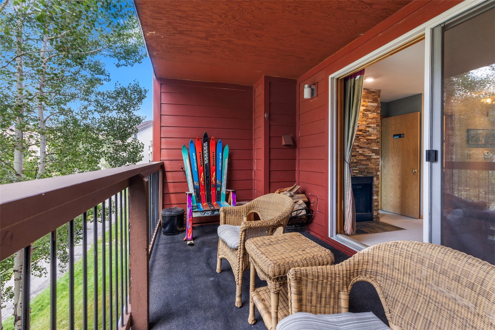 This covered balcony offers a cozy outdoor living space featuring a unique chair crafted from vintage skis and two wicker chairs with a matching side table. The space is finished with red siding and a dark floor, providing a warm, rustic atmosphere that overlooks a wooded area. A sliding glass door connects the balcony to an interior living area, where a stone-clad chimney breast is visible.
