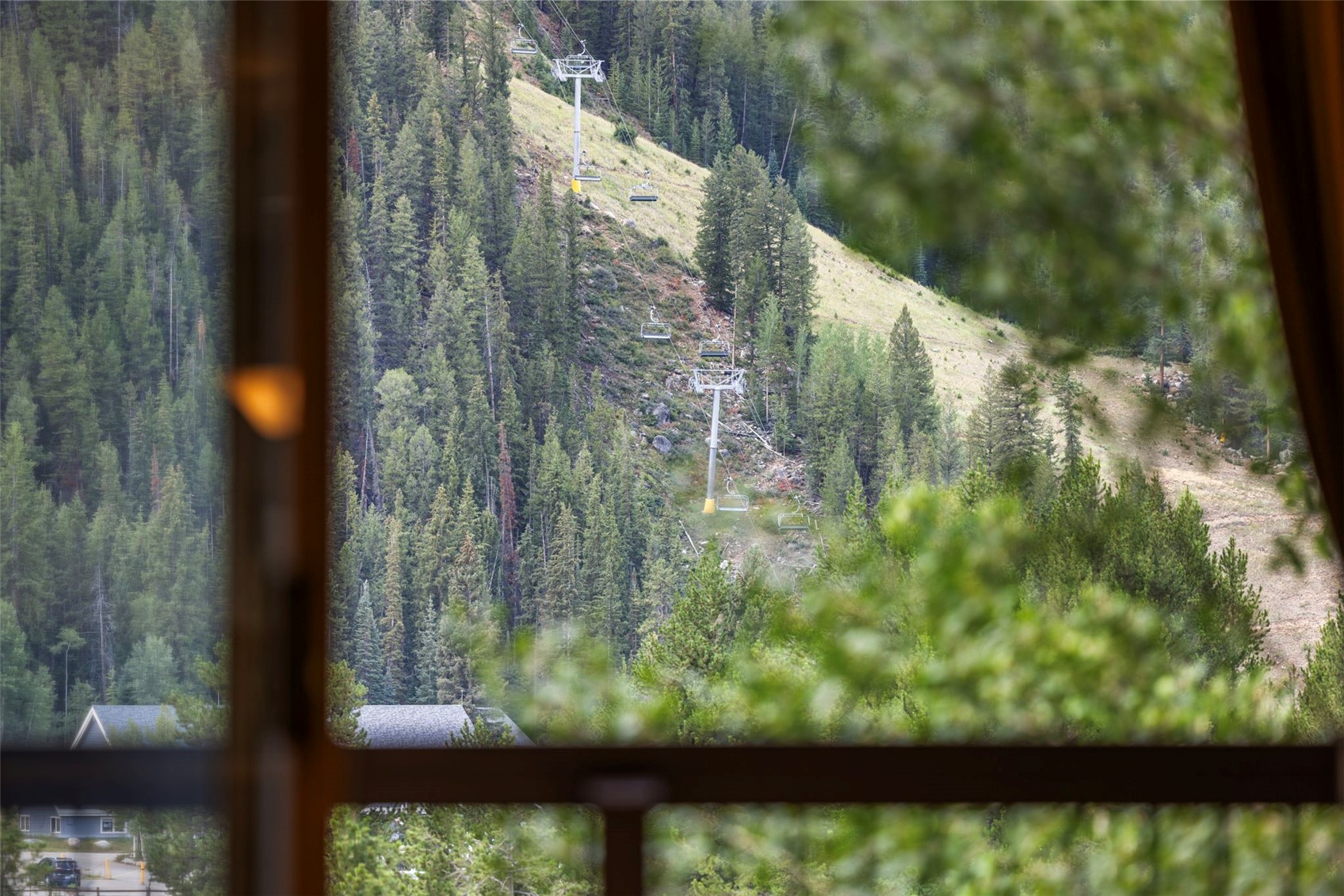 This image is captured from inside a home, looking out through a large window frame that divides the view into a grid. The perspective highlights a scenic mountain landscape featuring a ski lift ascending a forested slope. The foreground is slightly softened by out-of-focus foliage, creating a sense of depth and tranquility.