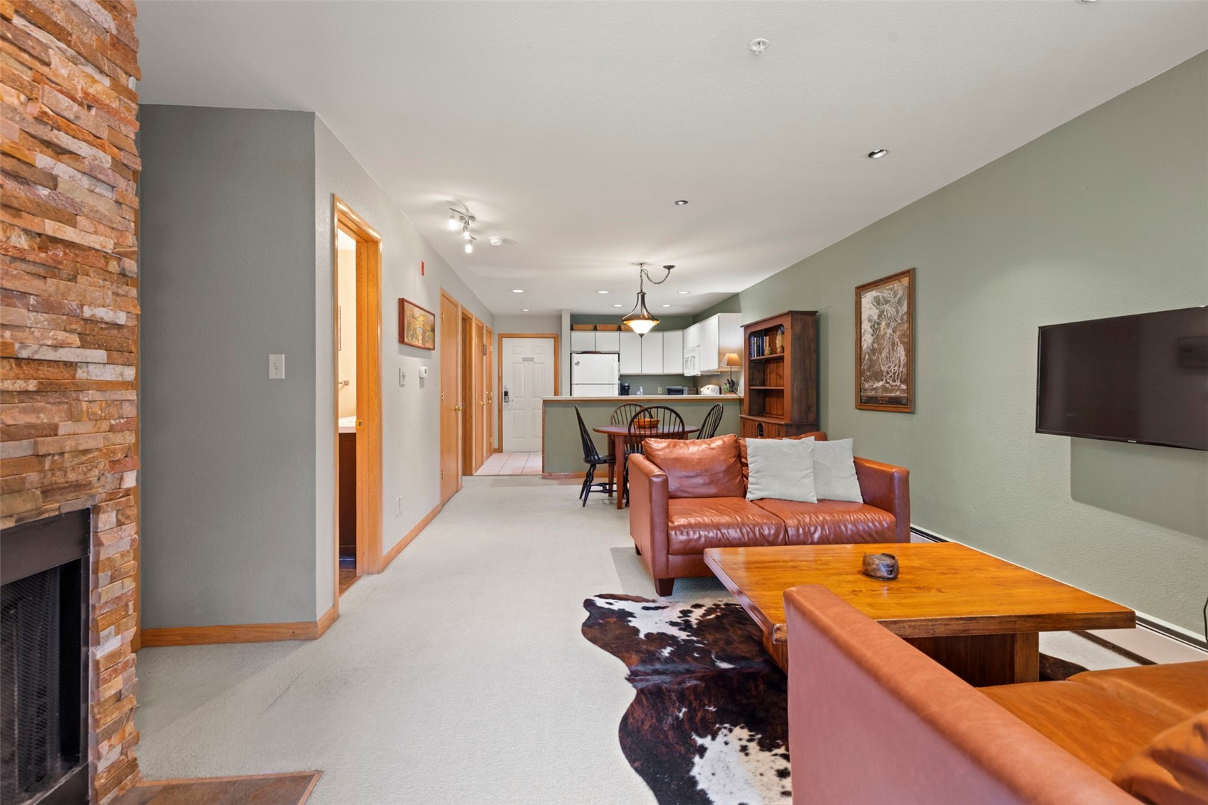 This open-concept living area features a warm, rustic aesthetic with a prominent stacked-stone chimney breast on the left and a cozy leather sofa facing a television. A cowhide rug sits beneath a wooden coffee table, while a dining area with a round table and chairs is positioned near the kitchen in the background. The space is finished with light-colored carpeting and sage green walls, creating a comfortable and inviting atmosphere.