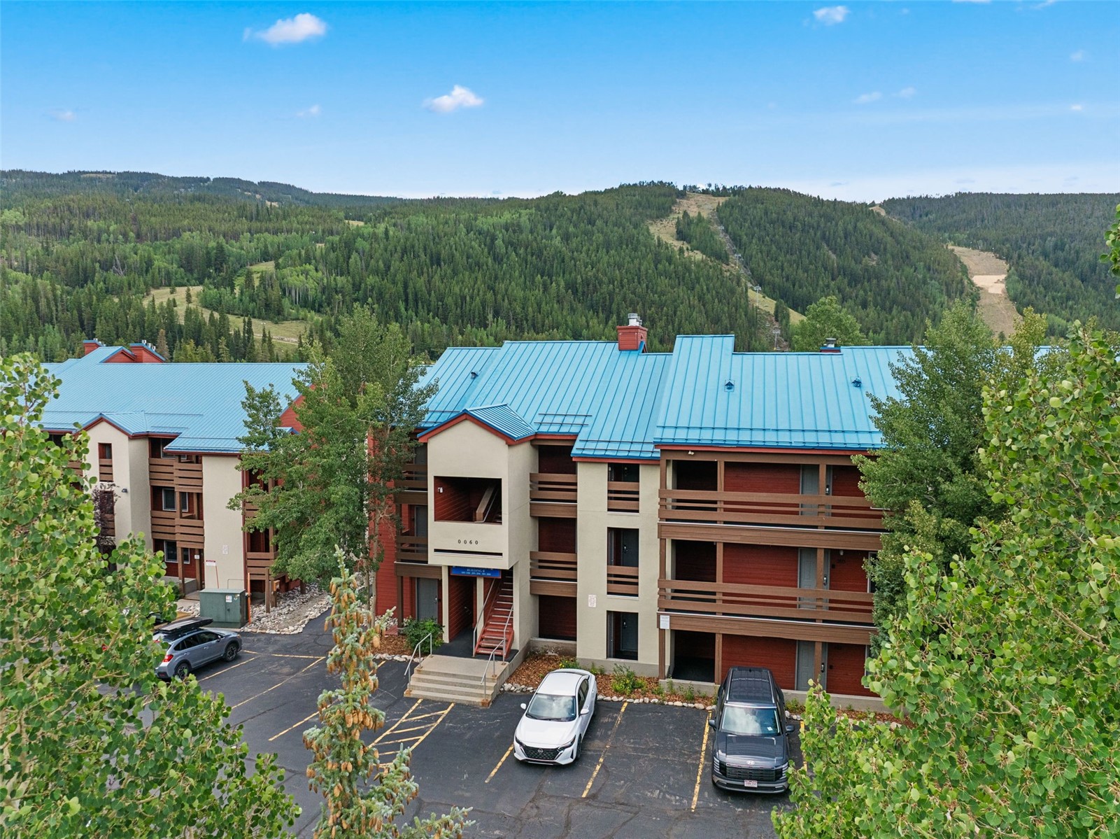 This aerial perspective captures a multi-story residential building with a distinctive blue metal roof, set against a backdrop of a lush, forested mountain slope. The structure features a combination of light-colored stucco and dark wood siding, with tiered balconies and a central entryway marked by the number 0060. A paved parking lot occupies the foreground, containing several parked vehicles and surrounded by mature green trees.
