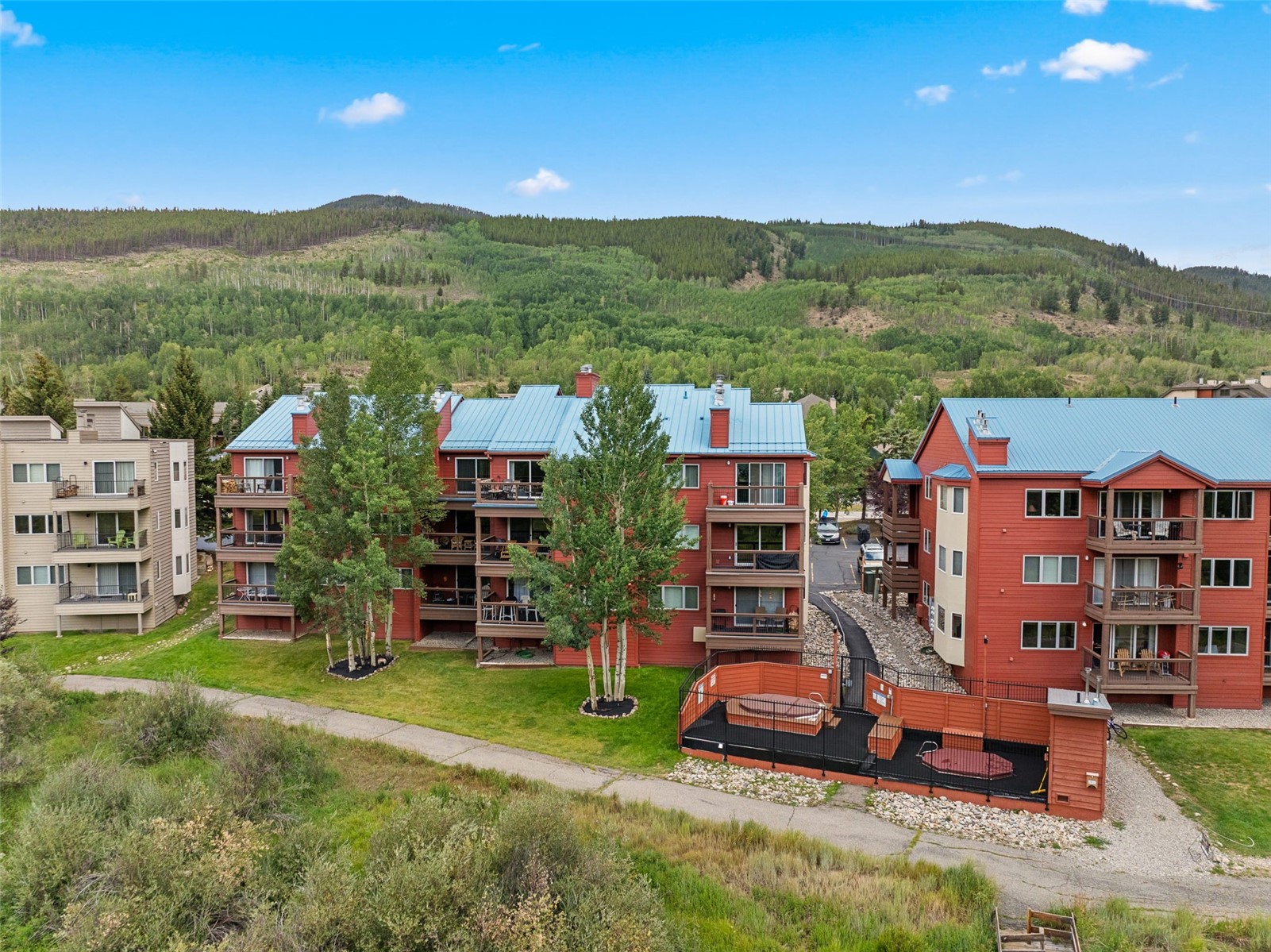 This aerial perspective captures a multi-story residential complex with red siding and blue metal roofing, nestled against a backdrop of lush, forested mountains. The property features multiple balconies, a paved pathway, and a fenced-in outdoor spa area with hot tubs. The scene conveys a serene, mountain-resort atmosphere, highlighting the integration of the buildings into the natural landscape.