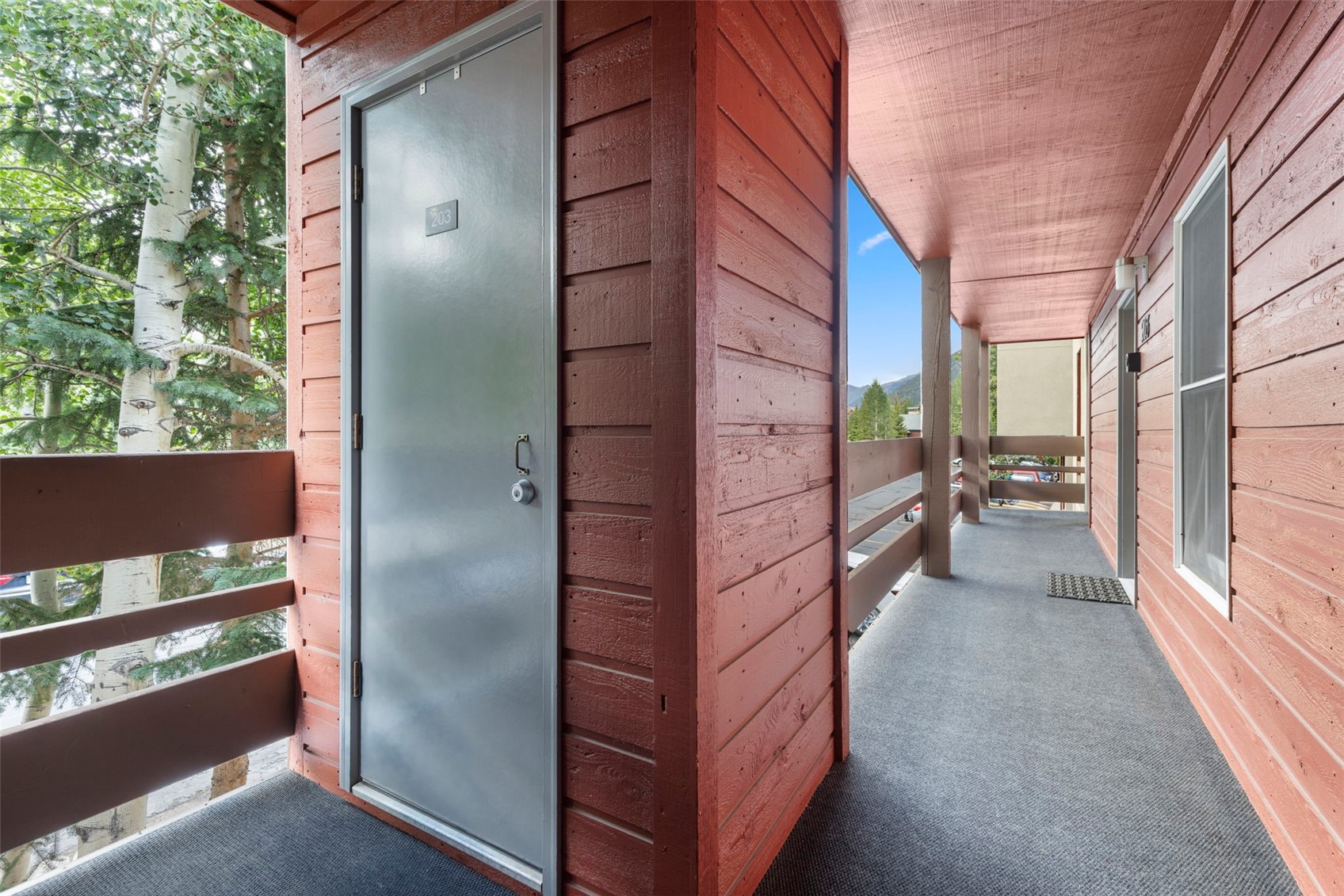 This image captures a covered outdoor balcony walkway featuring rustic, red-painted wood siding and a gray carpeted floor. The space includes a gray entry door marked with the number 203, a simple wooden railing overlooking the surrounding trees, and a window along the wall. The perspective looks down the length of the balcony, highlighting the transition from the shaded entryway to the bright, open view of the mountains in the distance.