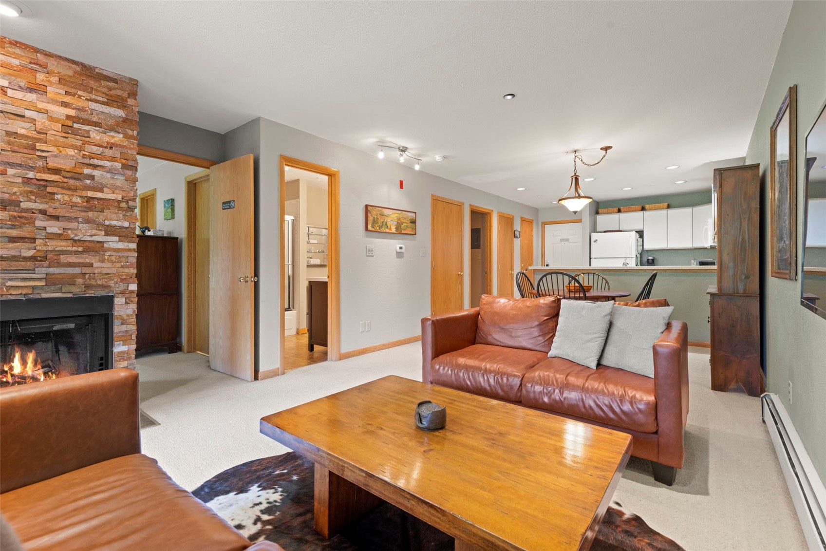 This open-concept living area features a cozy seating arrangement with brown leather sofas centered around a wooden coffee table on a cowhide rug. A prominent stacked-stone chimney breast anchors the room, while the space flows seamlessly into a dining area and kitchen in the background. The neutral walls and warm wood trim create a comfortable, inviting atmosphere suitable for a mountain or lodge-style home.