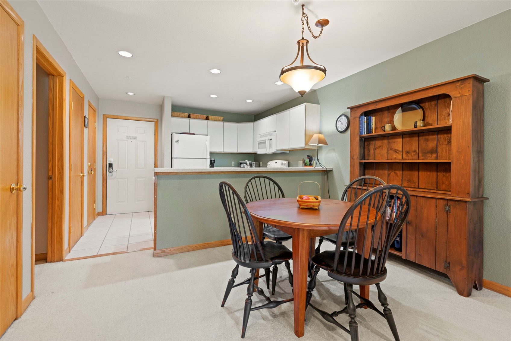 This dining area features a round wooden table surrounded by four black spindle-back chairs, positioned in front of a tall, rustic wooden hutch. The space is illuminated by a warm-toned pendant light fixture and opens directly into a kitchen with white cabinetry and a breakfast bar. The neutral wall color and carpeted flooring create a cozy, functional atmosphere suitable for casual dining.