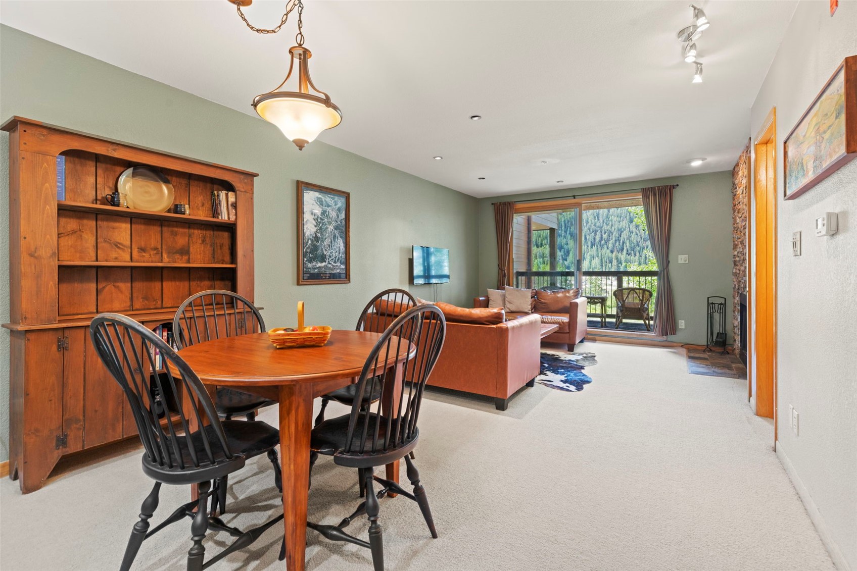 This open-concept living and dining area features a warm, rustic aesthetic with light green walls and neutral carpeting. The dining space includes a round wooden table with black spindle-back chairs, while the adjacent living area is furnished with a leather sofa and a cowhide rug. A large sliding glass door provides natural light and a view of the wooded exterior, complemented by a stone-clad chimney breast on the right side of the room.