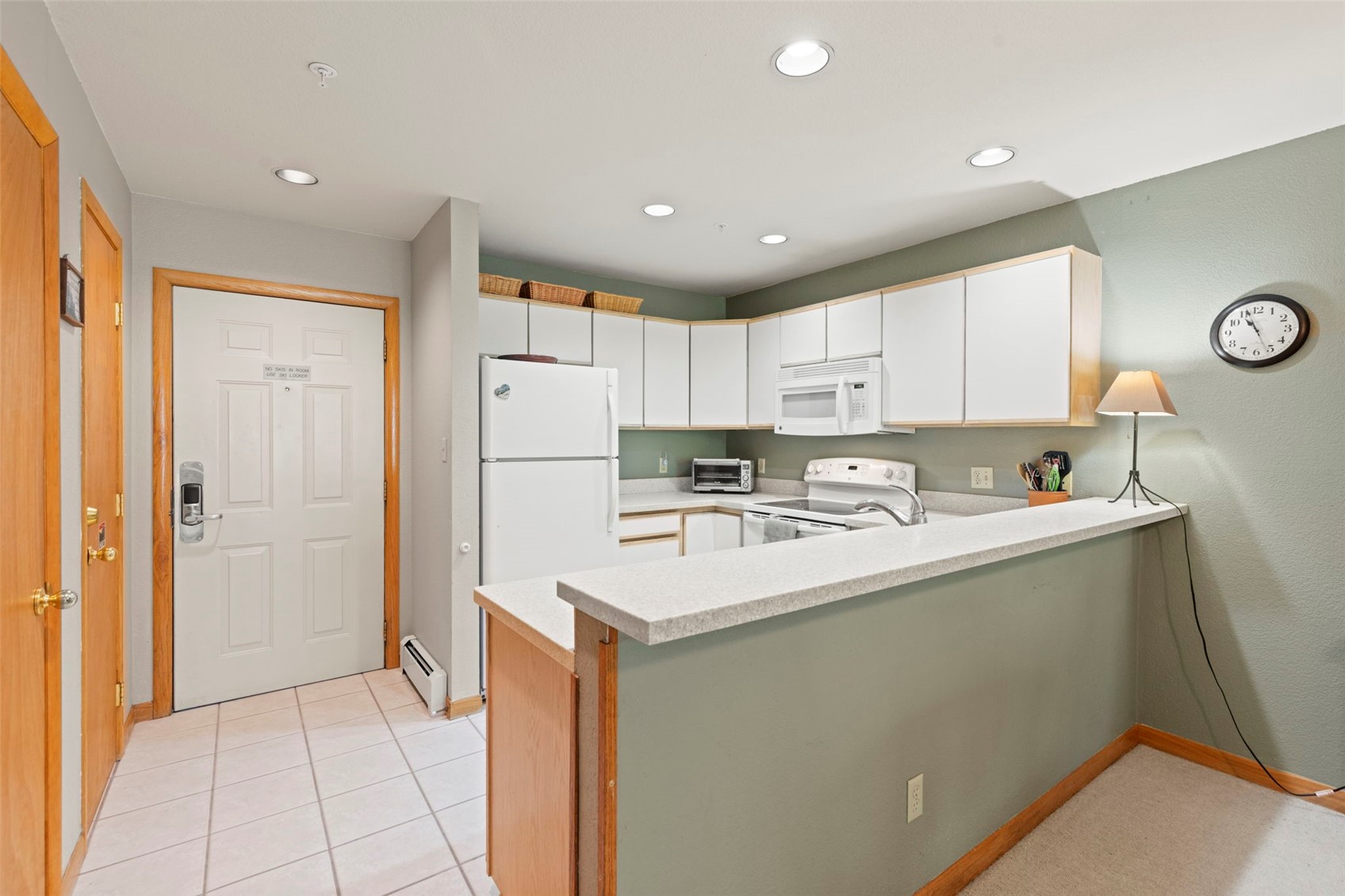 This kitchen features a functional U-shaped layout with white cabinetry, light-toned countertops, and a breakfast bar that opens into the adjacent living area. The space includes a white refrigerator, an electric range with an over-the-range microwave, and a neutral color palette with light green walls. A doorway leads to the entrance, and the room is illuminated by recessed ceiling lights, creating a bright and practical atmosphere.