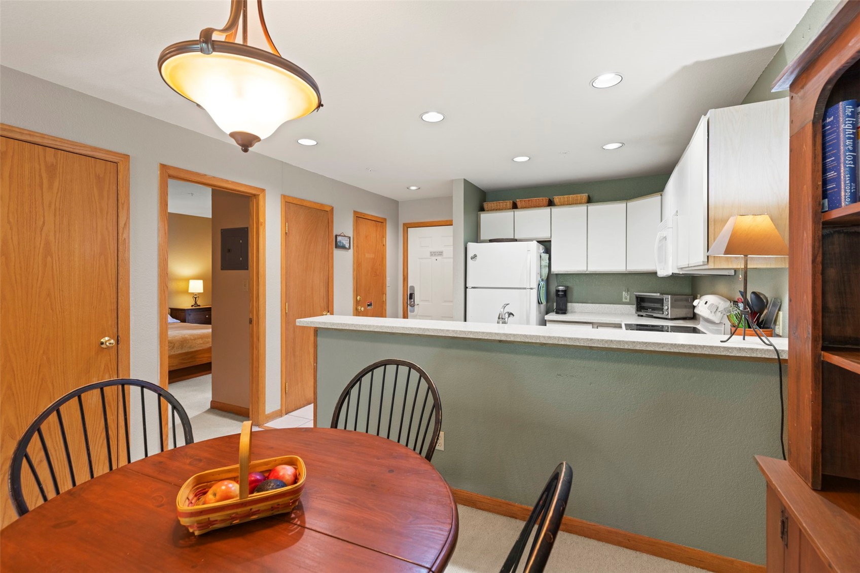 This interior view captures a functional kitchen and dining area, featuring a round wooden table in the foreground and a breakfast bar that separates the kitchen from the living space. The kitchen is equipped with white cabinetry, a white refrigerator, and a toaster oven, all set against a muted green wall. A warm, inviting atmosphere is created by the pendant light above the table and the lamp on the counter, with a glimpse into an adjacent bedroom through an open doorway.