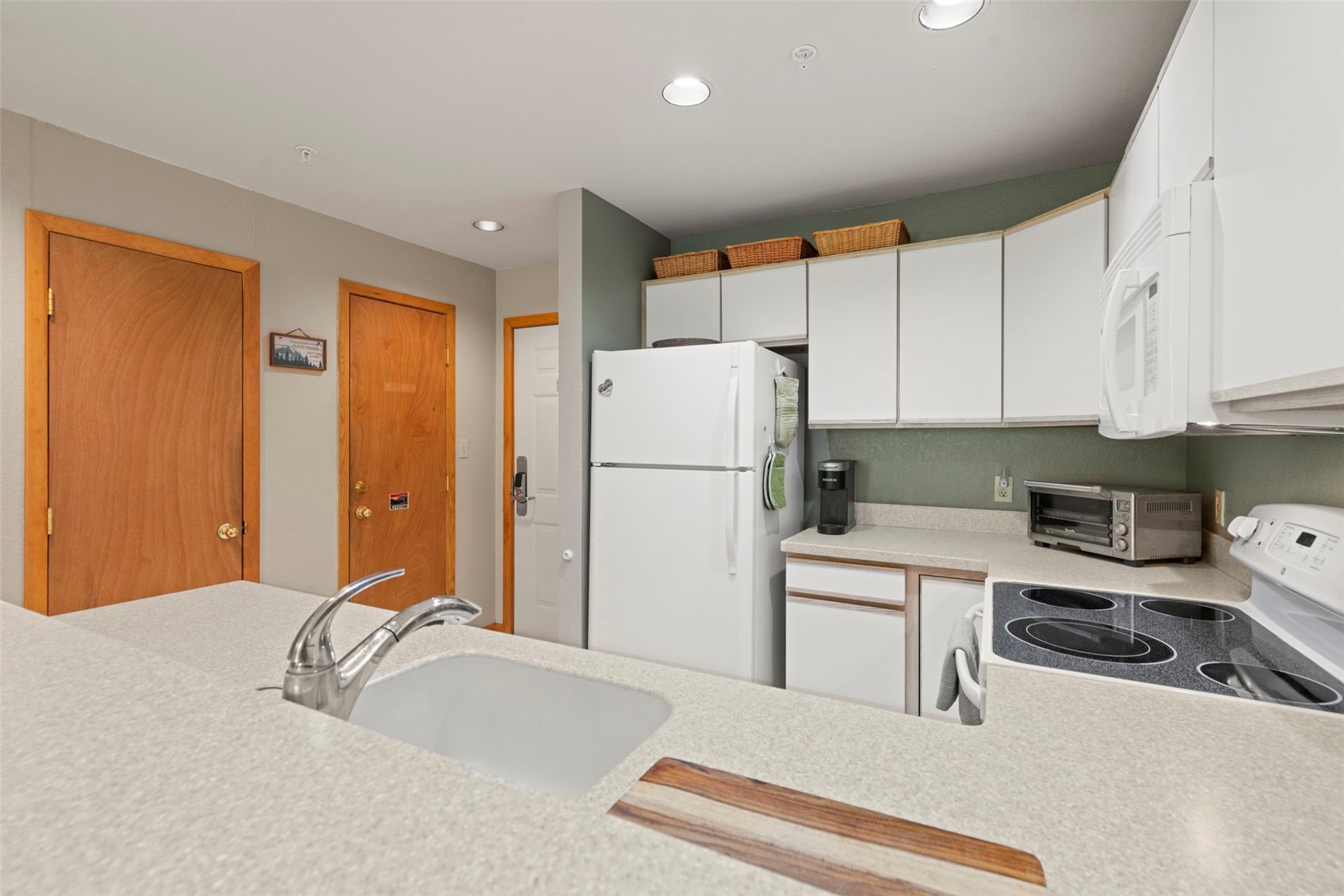 This kitchen features a functional layout with white cabinetry, a white refrigerator, and a smooth-top electric range. The space includes a peninsula with a sink and a light-colored countertop, providing a view into the adjacent living area. The walls are painted a muted green, and the room is accessed by multiple wooden doors, creating a practical and compact living environment.