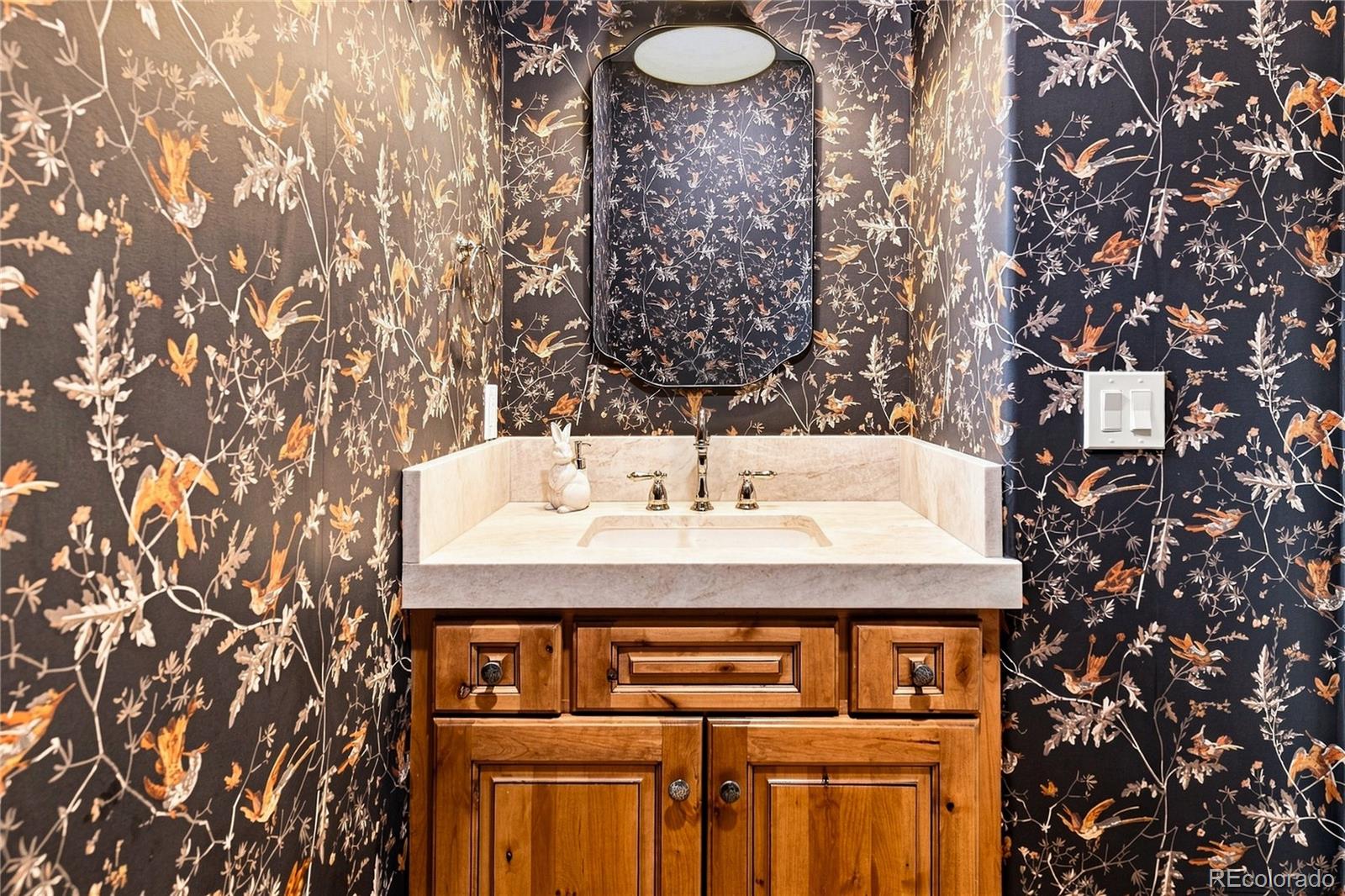 This powder room features bold, dark-toned wallpaper with a detailed bird and botanical pattern that creates a dramatic focal point. A light-colored stone vanity top with an integrated sink sits atop a warm-toned wood cabinet, providing a classic contrast to the dark walls. A decorative mirror is centered above the vanity, illuminated by a soft overhead light fixture, completing the sophisticated and moody aesthetic.