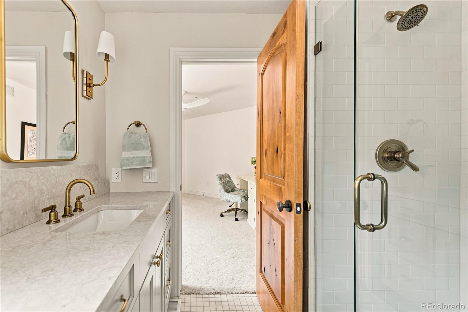 This bathroom features a vanity with a light-toned stone countertop, a gold-toned faucet, and a matching wall-mounted light fixture. A glass-enclosed shower with white subway tile is positioned to the right, while a wooden door stands slightly ajar, revealing a glimpse into an adjacent office space with carpeted flooring. The perspective captures the transition between the functional bathroom area and the quiet, bright workspace beyond.