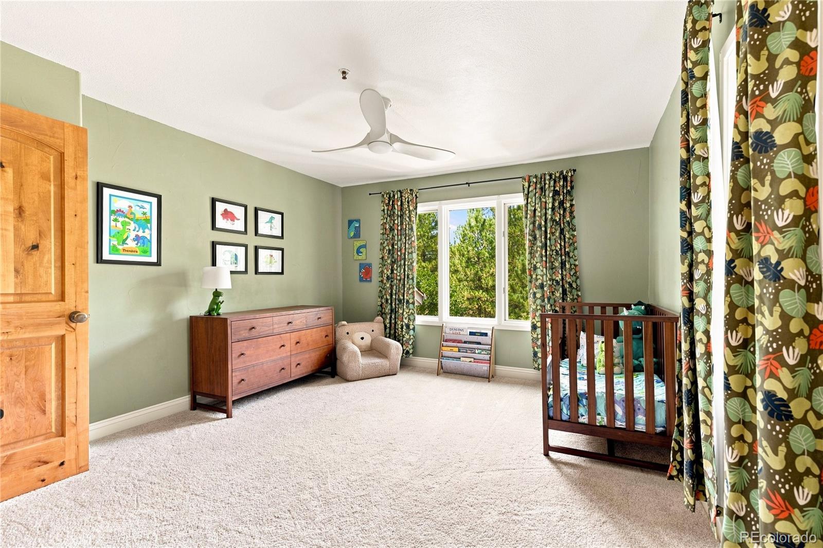 This charming nursery features soft green walls, neutral carpeting, and a wooden crib, creating a warm and inviting atmosphere. A wooden dresser, a small bear-shaped armchair, and a book rack provide functional storage and seating, while patterned curtains frame the large window overlooking trees. The room is decorated with dinosaur-themed artwork, making it a playful and well-appointed space for a child.