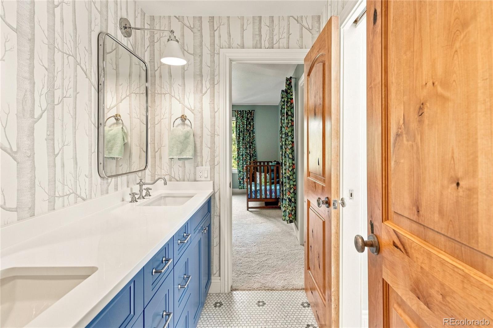 This bathroom features a vibrant blue vanity with a white countertop and dual sinks, set against whimsical tree-patterned wallpaper. A rectangular mirror with a rounded frame hangs above the vanity, complemented by a wall-mounted light fixture. The room leads directly into a bedroom visible through the doorway, showcasing a cohesive transition between spaces.