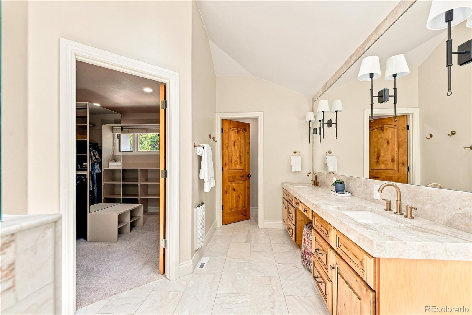 This primary bathroom features a dual-sink vanity with light wood cabinetry and a neutral stone countertop, complemented by a large wall-to-wall mirror and elegant wall-mounted light fixtures. The space includes light-colored tile flooring and leads into a walk-in closet through an open doorway, while a separate wooden door provides access to another area. The overall aesthetic is warm and traditional, offering a functional and well-lit layout.