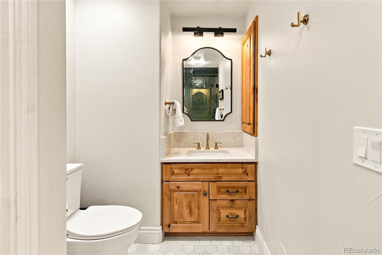This compact powder room features a rustic wood vanity with a stone countertop and a decorative arched mirror. The space is accented by brass-toned hardware, including a faucet and wall-mounted hooks, set against light-colored walls and a hexagonal tile floor. A simple light fixture above the mirror and a white toilet complete this functional and well-lit bathroom.