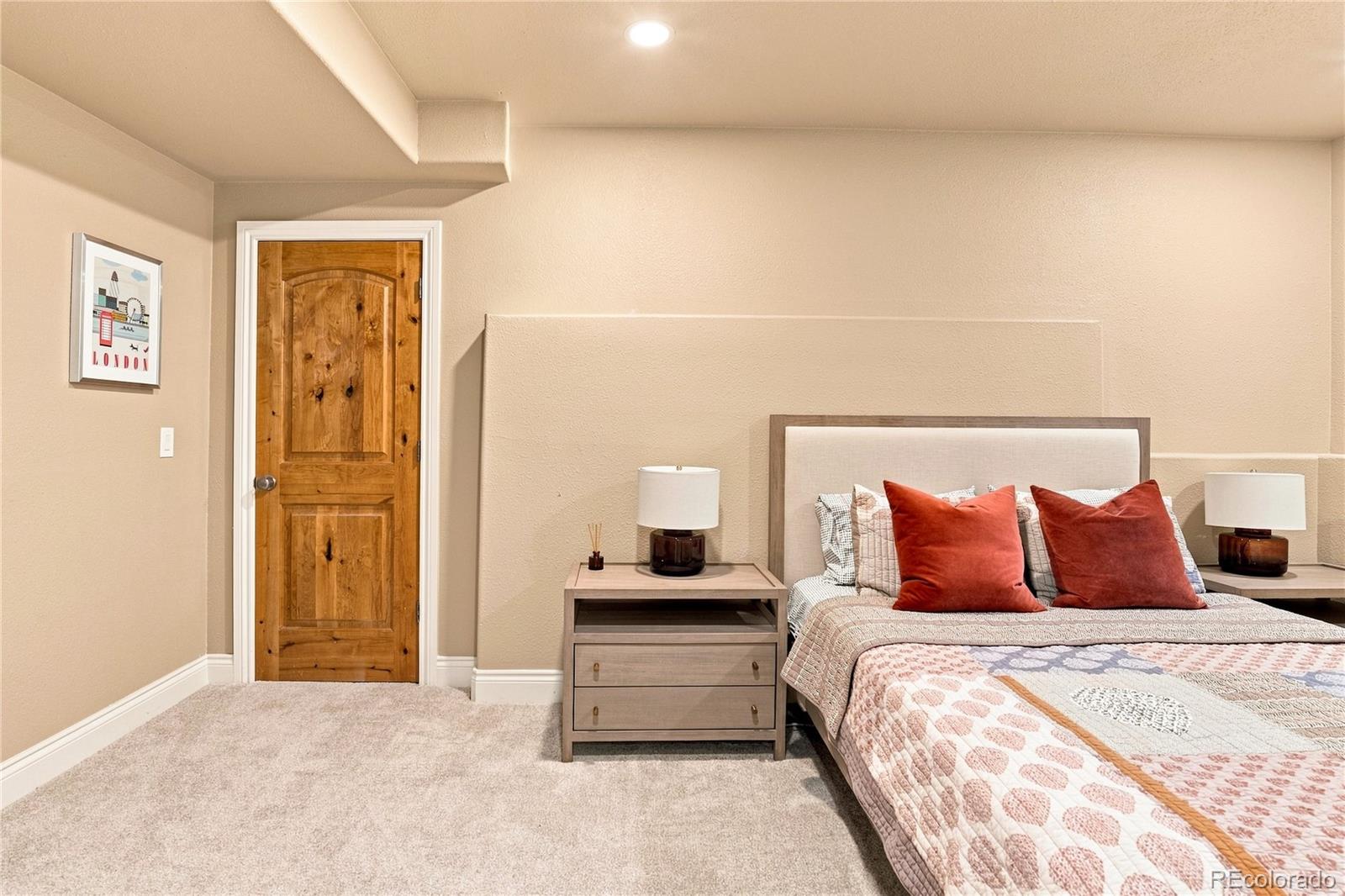 This cozy bedroom features a neutral color palette with a light-toned upholstered headboard and matching nightstands. A rustic, knotty wood door adds a touch of warmth to the space, while a framed London-themed print provides a decorative accent on the wall. The room is carpeted and includes a patterned quilt with rust-colored throw pillows, creating a comfortable and inviting atmosphere.