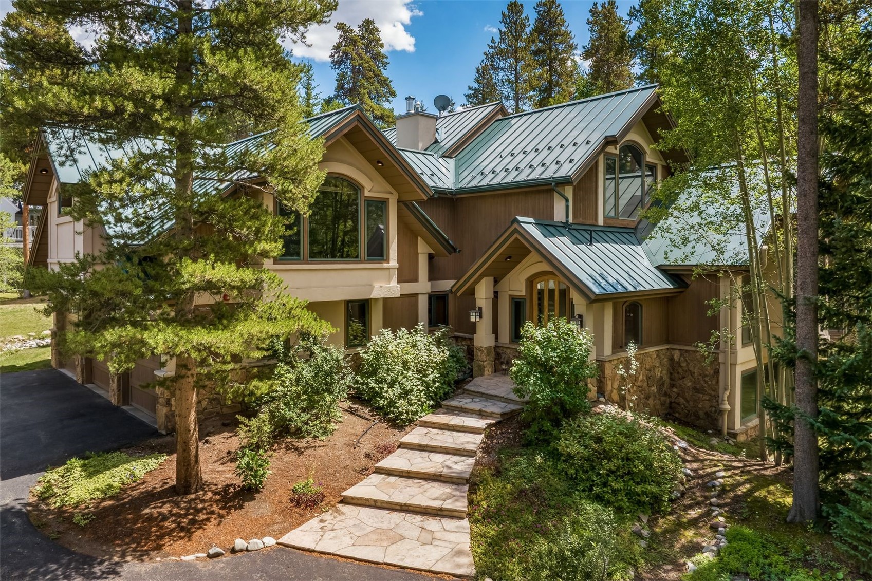 This mountain-style residence features a multi-gabled roofline with green metal roofing and a mix of wood siding and stone masonry. A stone-paved walkway leads through a landscaped front yard to the covered entryway, framed by mature pine and aspen trees. The home's architectural design emphasizes a rustic, woodland aesthetic that blends seamlessly into the surrounding forest environment.