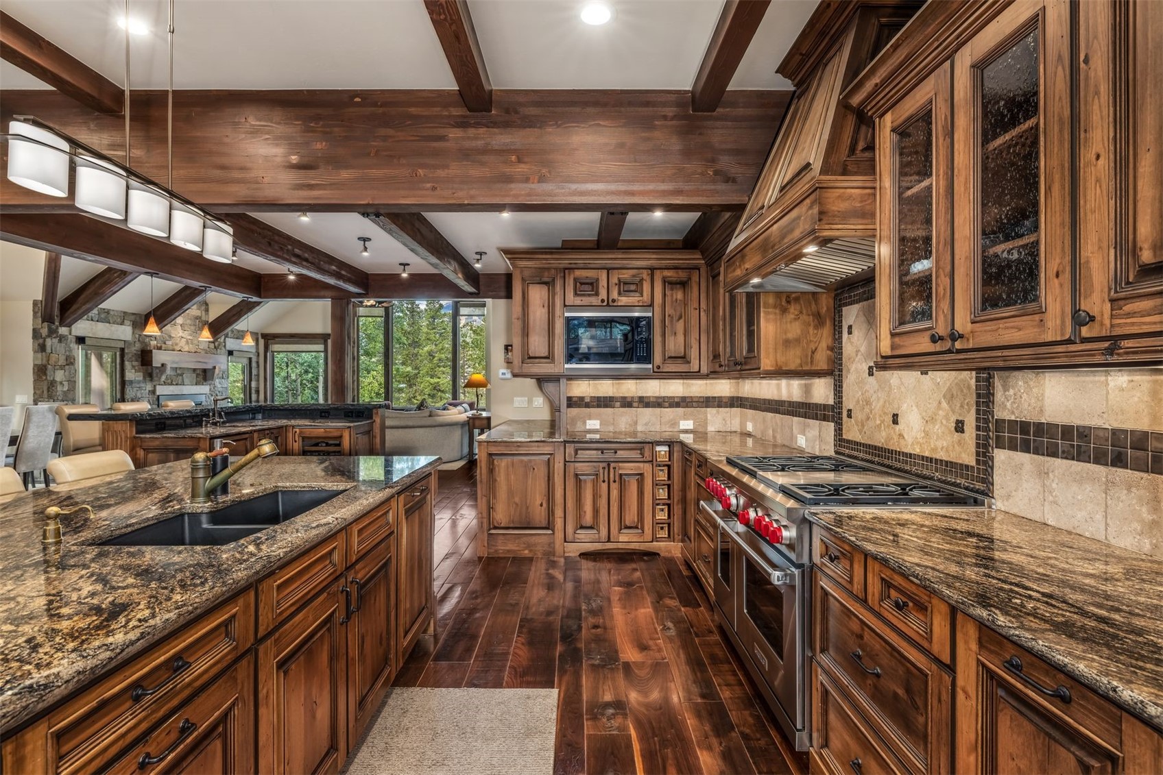 This rustic-style kitchen features rich, dark wood cabinetry, exposed timber ceiling beams, and expansive stone countertops. A large central island with a dark sink anchors the space, while professional-grade stainless steel appliances and a decorative stone backsplash add a touch of luxury. The open-concept layout flows seamlessly into a living area, creating a warm and inviting atmosphere with views of the surrounding greenery through large windows.