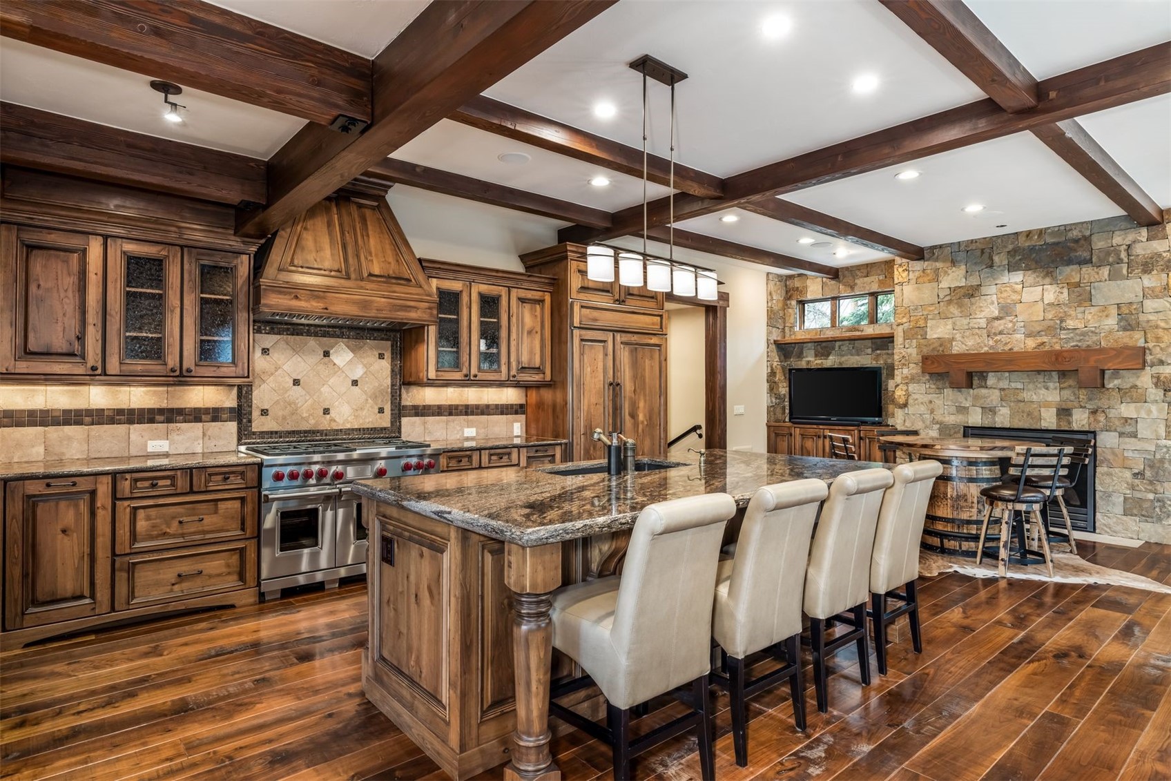 This rustic-style kitchen features rich, dark wood cabinetry, a large central island with seating for four, and a professional-grade range with a custom wood hood. The space is defined by exposed ceiling beams and a prominent stone-clad chimney breast that houses a television and fireplace. The warm, inviting atmosphere is enhanced by polished wood flooring and a multi-light pendant fixture hanging over the island.