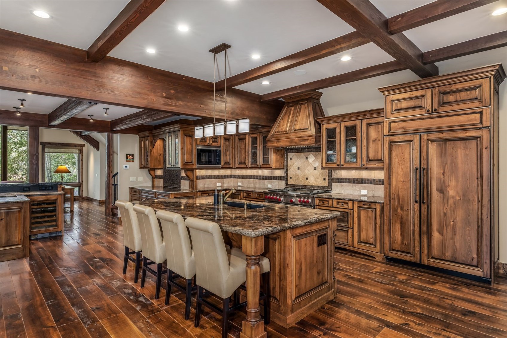 This rustic-style kitchen features extensive dark wood cabinetry, exposed ceiling beams, and a large central island with a stone countertop and seating for four. The space is equipped with professional-grade appliances, including a gas range with a custom wood hood and a built-in refrigerator, all set against a backdrop of warm wood flooring. The perspective captures the expansive layout, highlighting the blend of traditional craftsmanship and modern functionality.