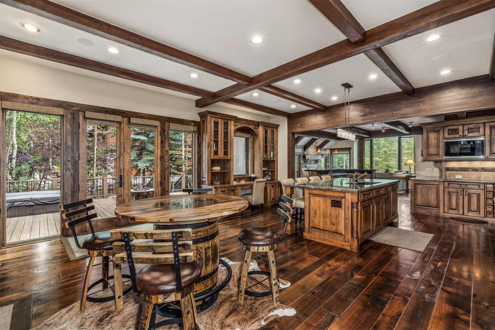 This rustic-style kitchen and dining area features rich wood cabinetry, exposed ceiling beams, and dark wood flooring. A unique barrel-base table sits on a cowhide rug in the foreground, while the kitchen island provides ample counter space and seating. Large glass doors open to an outdoor deck, creating a seamless transition between the indoor living space and the natural surroundings.