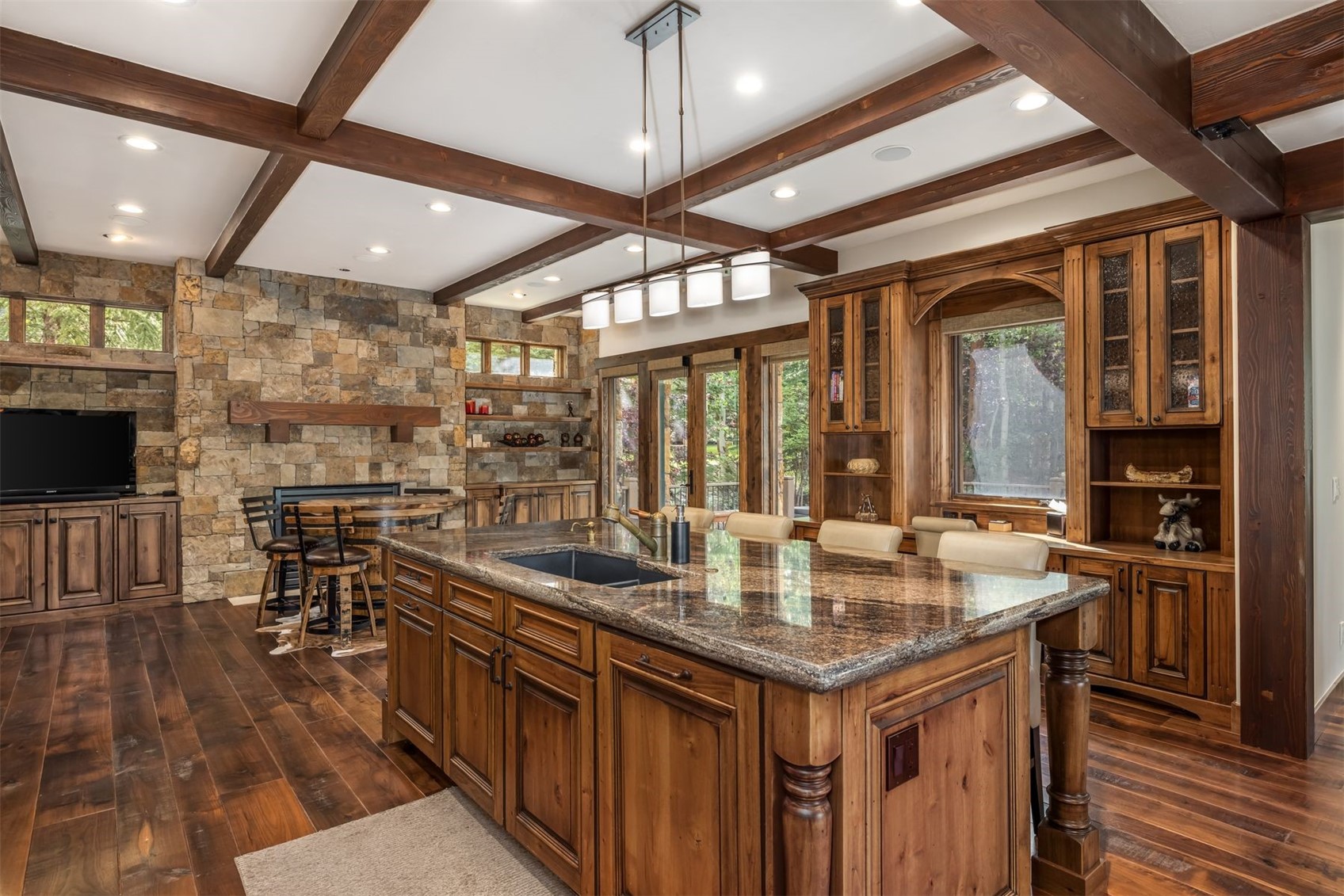 This rustic-style kitchen features a large central island with a dark stone countertop and a built-in sink, complemented by warm-toned wood cabinetry throughout. A prominent stone-clad chimney breast serves as a focal point, housing a fireplace and a television, while exposed wooden ceiling beams add architectural character. The space is illuminated by a multi-light pendant fixture over the island and recessed lighting, creating a cozy and inviting atmosphere.
