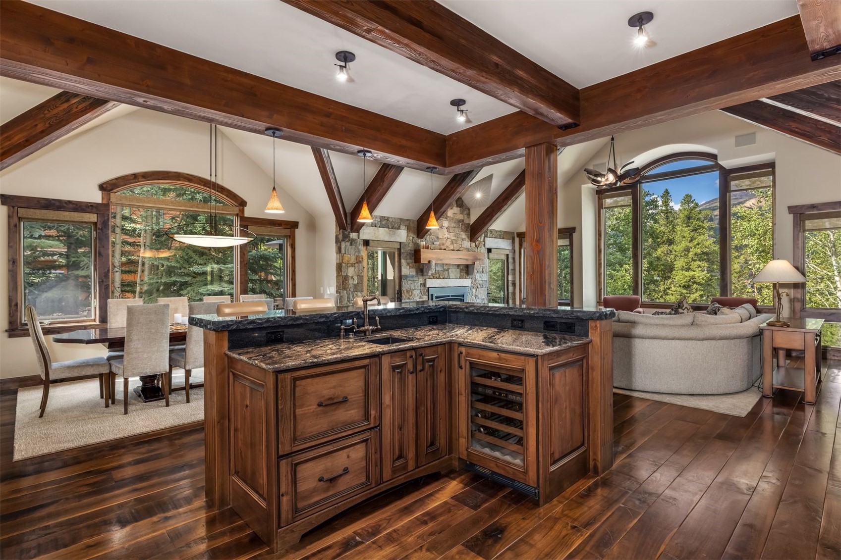 This open-concept kitchen features a central island with a dark stone countertop, a built-in wine cooler, and rich wood cabinetry. The space is defined by exposed timber beams and warm wood flooring, seamlessly connecting to a dining area on the left and a cozy living room on the right. Large windows throughout the space provide abundant natural light and scenic views of the surrounding forest.