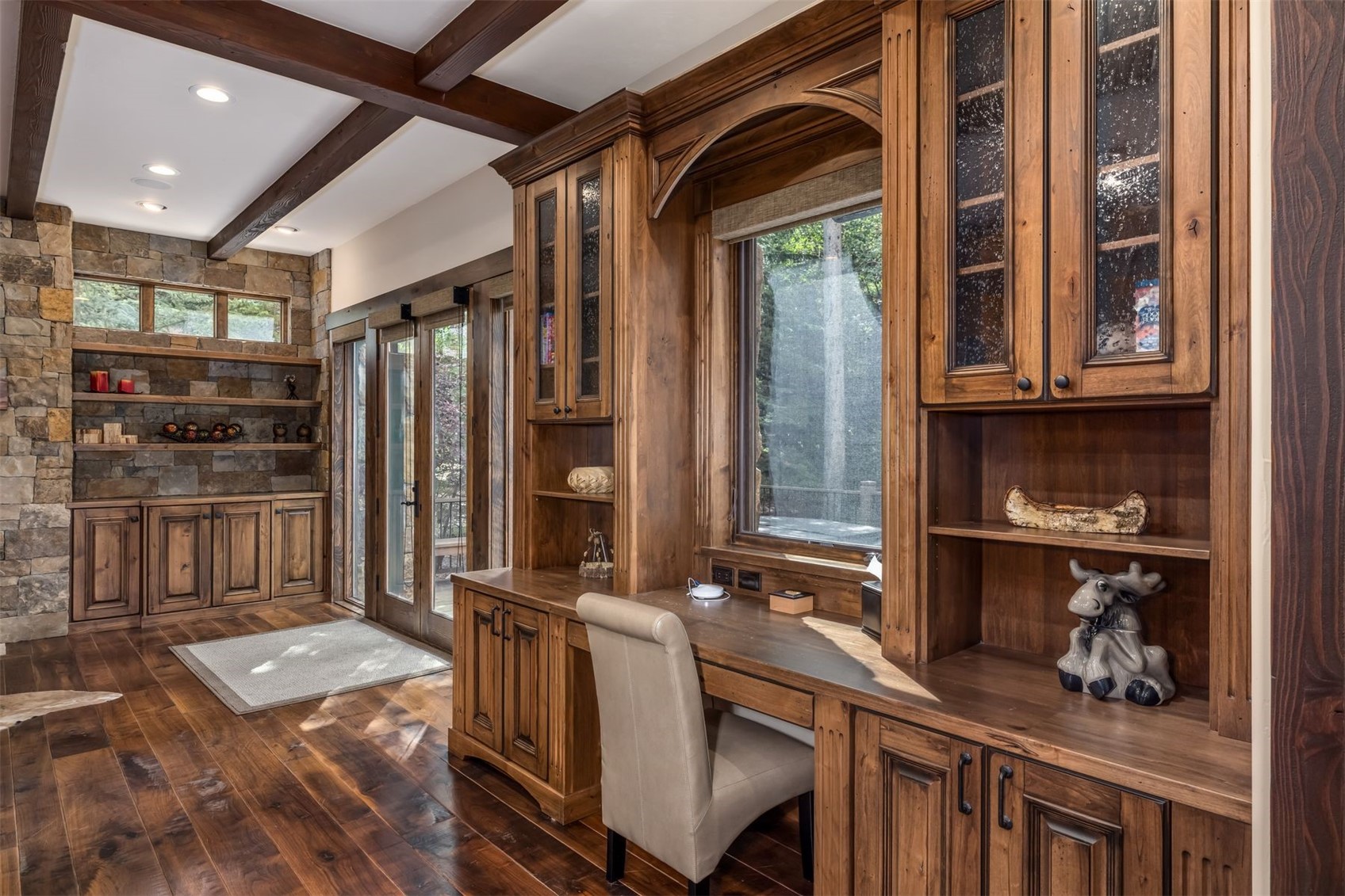 This rustic home office features extensive custom wood cabinetry with glass-front upper storage and a built-in desk area centered under a window. To the left, a stone-clad wall incorporates open shelving and lower cabinets, complemented by dark wood ceiling beams and hardwood flooring. The space offers a warm, sophisticated atmosphere with a seamless transition to an adjacent outdoor area through glass-paned doors.