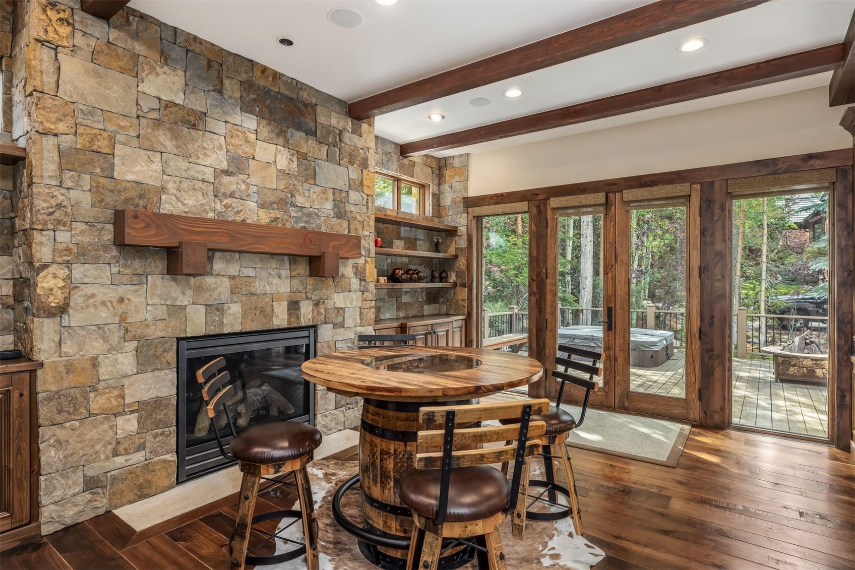 This rustic dining area features a unique table crafted from a wooden barrel base and a circular top, surrounded by matching stools with leather seats. A prominent stone-clad chimney breast serves as the focal point, complete with a heavy timber mantel and an integrated fireplace. The space is connected to an outdoor deck through a series of glass-paned doors, which provide a view of the surrounding forest and a private hot tub.