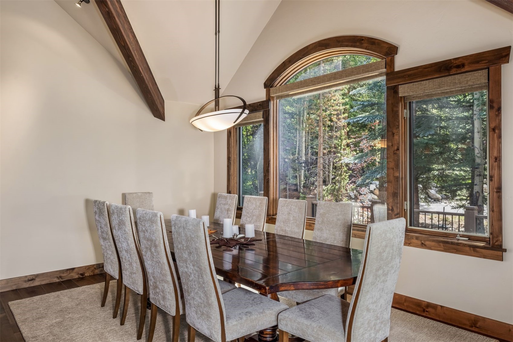 This elegant dining room features a large, dark wood table surrounded by ten upholstered chairs with a light-colored, textured fabric. A prominent arched window with dark wood trim provides a scenic view of the surrounding forest, while a modern pendant light hangs above the table. The room is accented by a vaulted ceiling with exposed wood beams, creating a warm and inviting atmosphere.