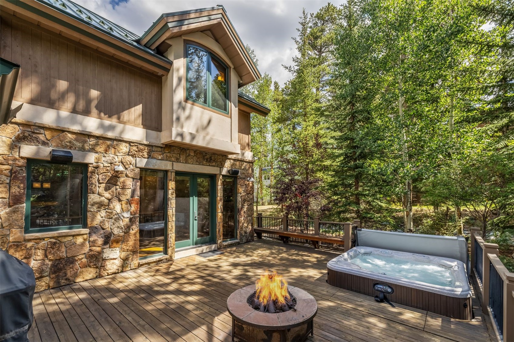 This spacious wooden deck features a stone-clad fire pit as a central gathering point, complemented by a private hot tub situated to the right. The exterior of the home showcases a blend of natural stone masonry and warm-toned wood siding, creating a rustic mountain aesthetic. Tall windows and glass doors provide a seamless transition between the indoor living space and the serene, tree-lined outdoor environment.