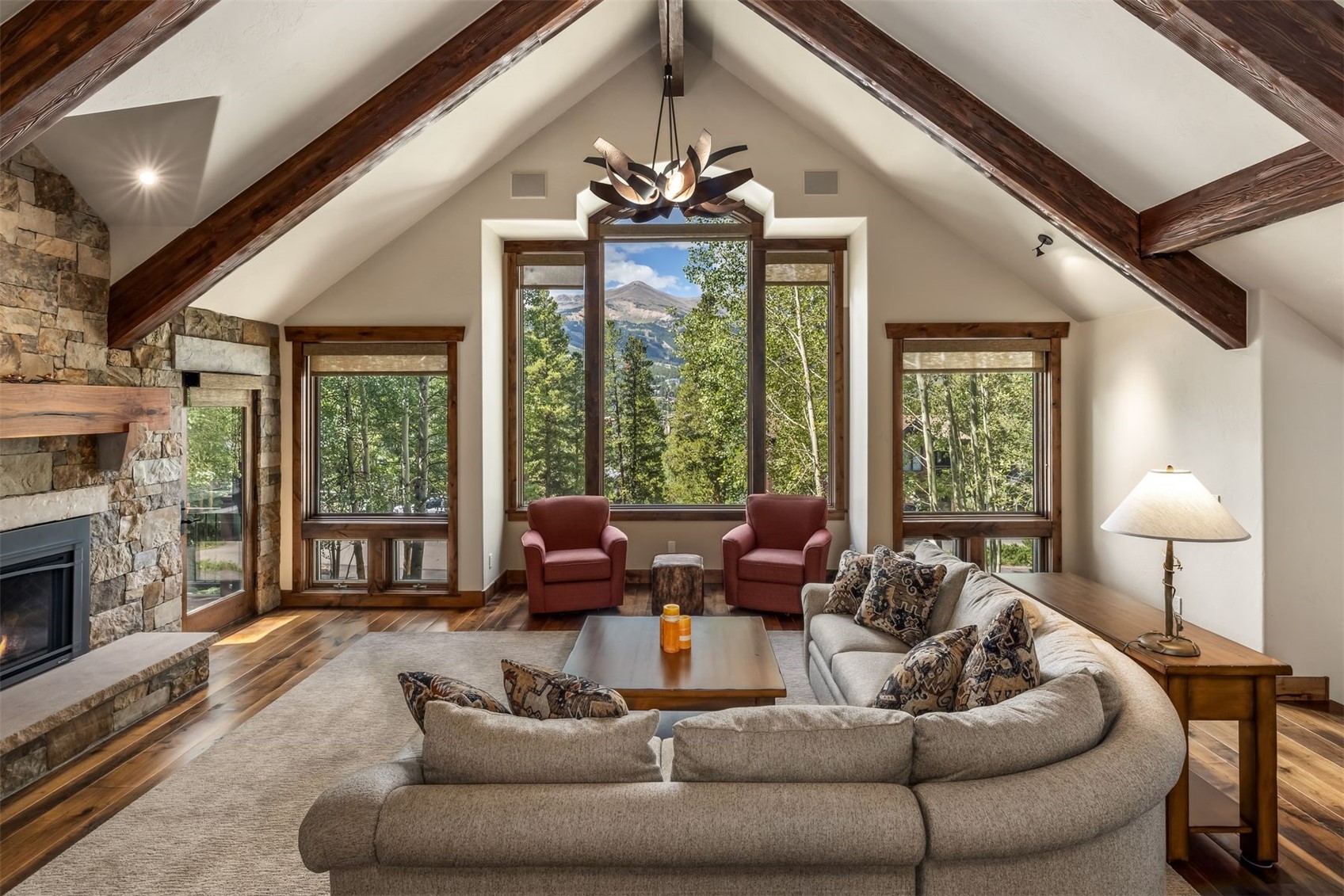 This spacious living room features a vaulted ceiling with exposed wooden beams and a prominent stone-clad chimney breast. A large sectional sofa and two armchairs are arranged around a central coffee table, creating a cozy seating area that faces a scenic mountain view through expansive windows. The room is anchored by a neutral area rug over hardwood flooring, with a decorative chandelier hanging from the peak of the ceiling.