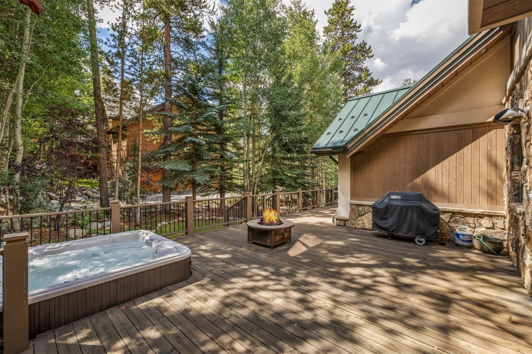 This spacious wooden deck offers a serene outdoor living space nestled among mature pine and aspen trees. The area features a built-in hot tub, a central fire pit with a flickering flame, and a covered grill station, all enclosed by a sturdy railing. The rustic architecture, characterized by wood siding and a green metal roof, creates a cozy mountain retreat atmosphere.
