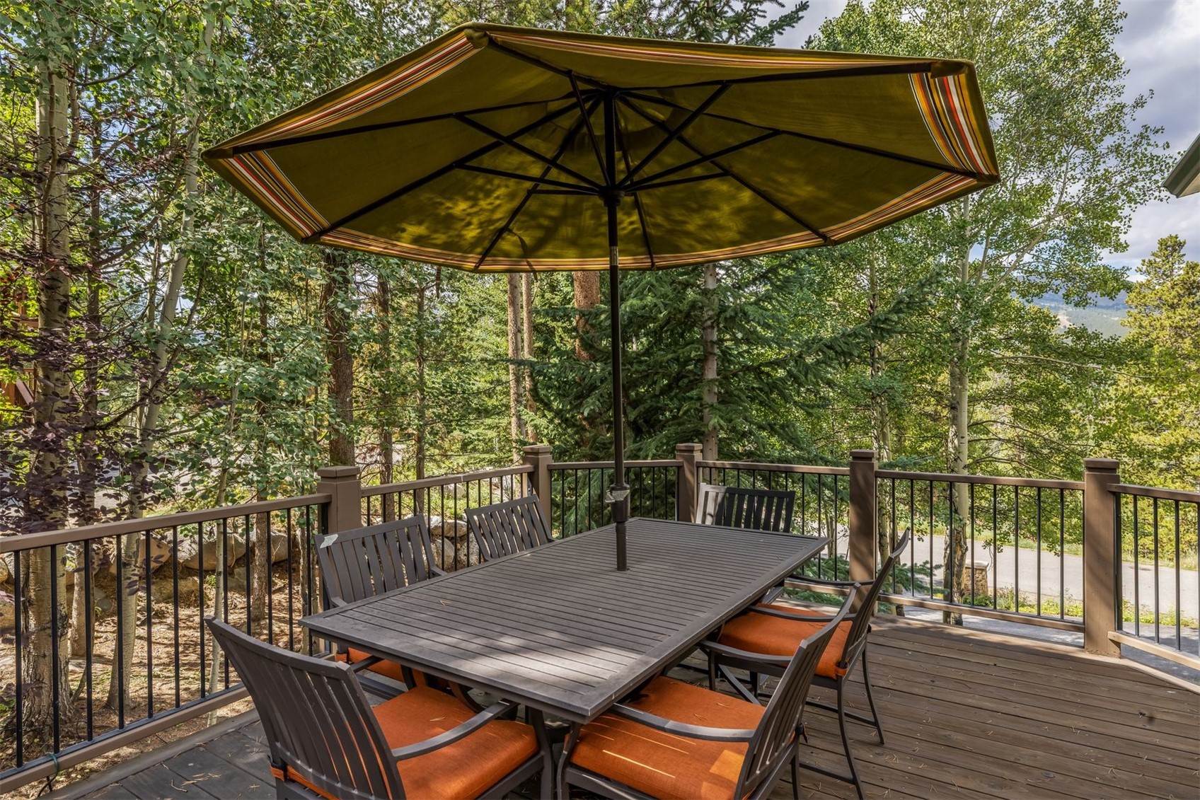 This elevated wooden deck features a dark-toned dining table set with six chairs, each equipped with vibrant orange cushions. A large, olive-green umbrella with striped trim provides shade, while a dark metal railing surrounds the perimeter. The deck is nestled among lush, mature trees, offering a serene and private outdoor dining space with a natural, wooded backdrop.