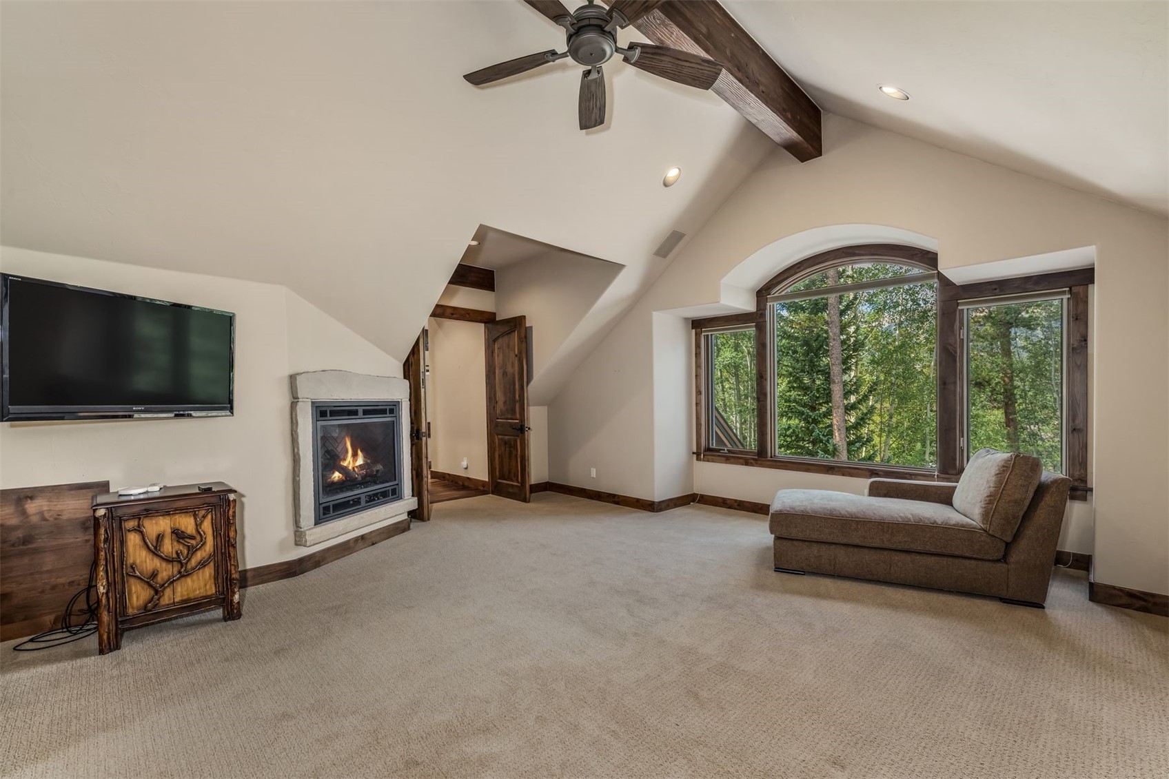This cozy living space features a vaulted ceiling with exposed wooden beams and a ceiling fan, creating a rustic, mountain-lodge atmosphere. A stone-surround fireplace provides a focal point, while a large arched window offers a serene view of the surrounding forest. The room is furnished with a comfortable chaise lounge and a decorative wooden cabinet, all set against a neutral carpeted floor.