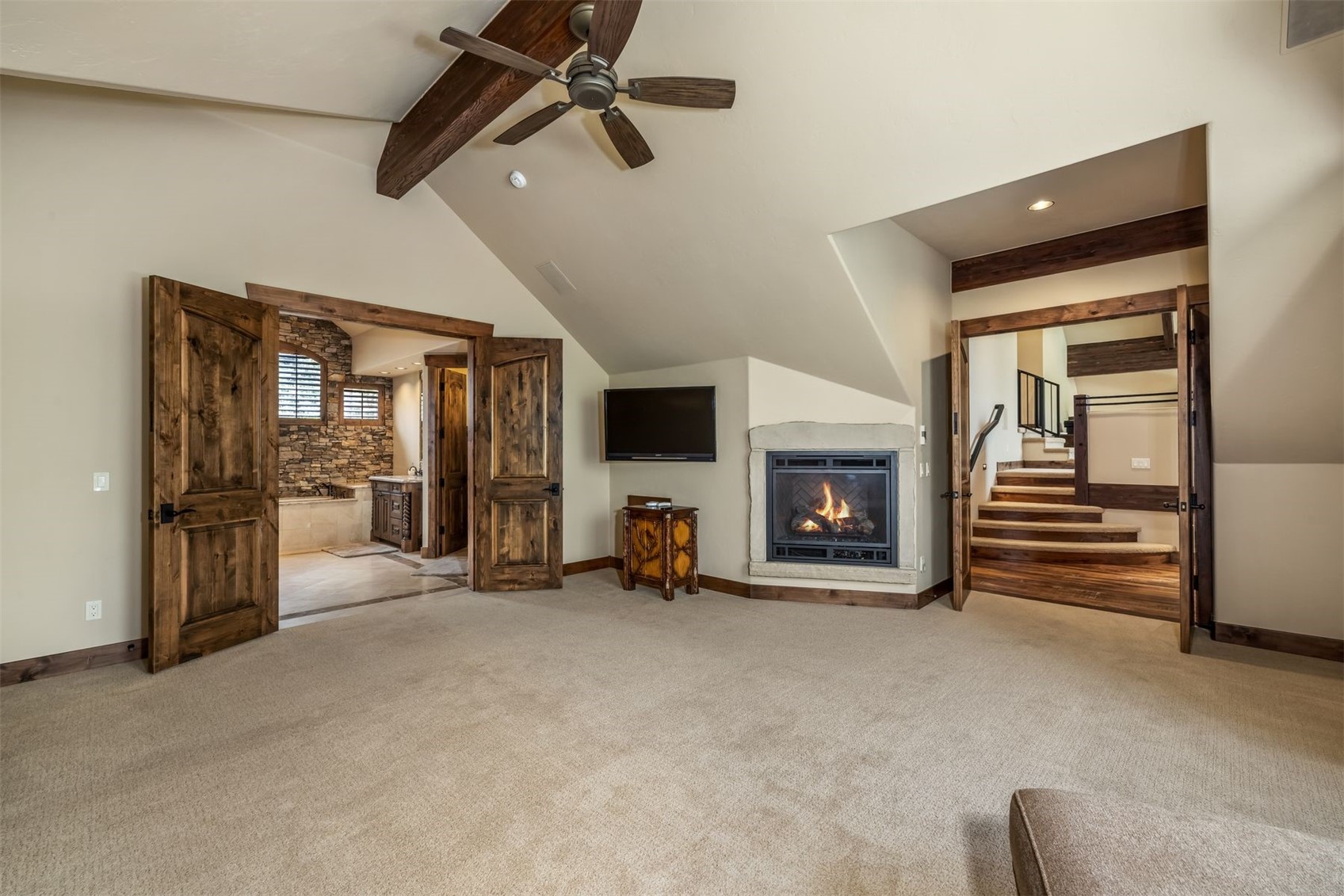This spacious primary bedroom features a vaulted ceiling with exposed wooden beams and a ceiling fan, creating a rustic and cozy atmosphere. The room includes a stone-clad fireplace, a wall-mounted television, and double wooden doors leading to an adjoining bathroom, while a separate arched doorway reveals a staircase. The neutral carpet and warm wall tones provide a comfortable, inviting aesthetic suitable for a master suite.