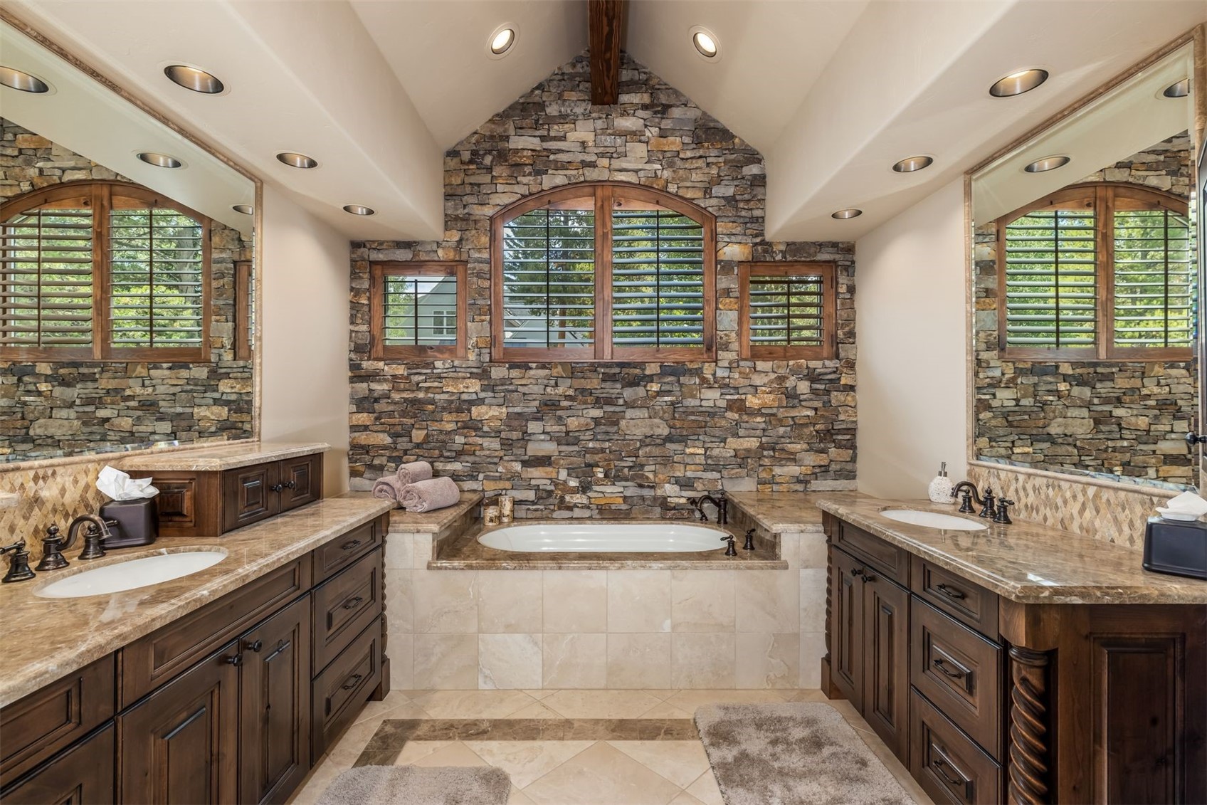 This primary bathroom features a symmetrical layout with dual vanities flanking a central soaking tub set against a prominent stone-clad wall. The space is defined by dark wood cabinetry, neutral stone countertops, and a vaulted ceiling with recessed lighting. The combination of the textured stone wall and arched windows creates a warm, rustic, and luxurious atmosphere.