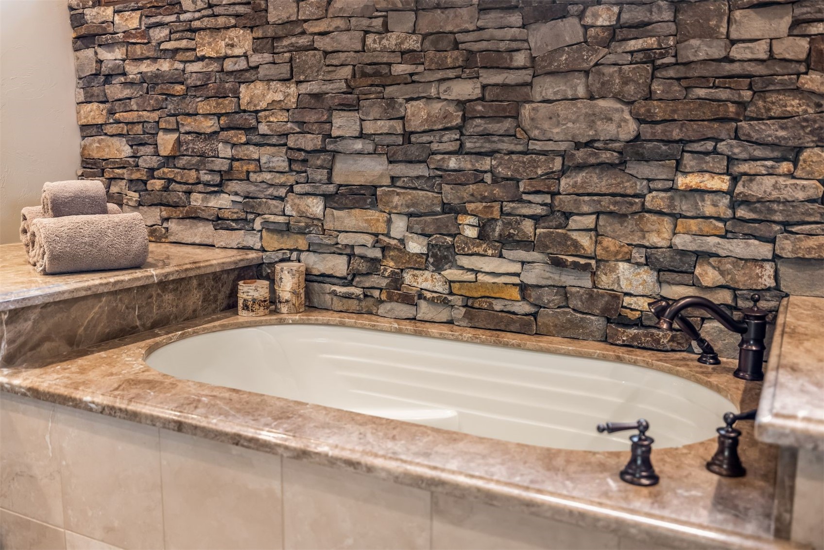 This primary bathroom features a deep soaking tub set within a stone-topped deck, accented by a rustic stacked-stone wall backdrop. The space includes dark bronze-toned fixtures and a pair of decorative birch-patterned canisters, creating a warm, spa-like atmosphere. The perspective is a close-up, eye-level shot focusing on the tub's luxurious design and the textured stone wall behind it.