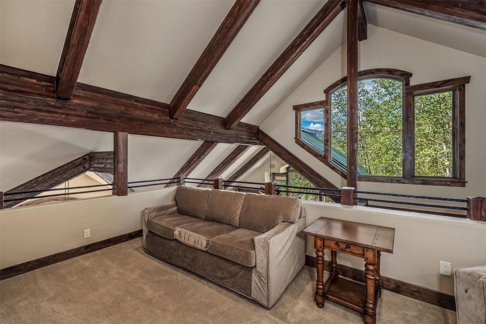 This cozy loft space features a vaulted ceiling with exposed dark wood beams and a neutral-toned sofa positioned against a low wall. A rustic wooden side table sits beside the sofa, while large, uniquely shaped windows provide a view of the surrounding trees and distant mountains. The room is carpeted and maintains an open, airy feel, overlooking the level below through a metal railing.