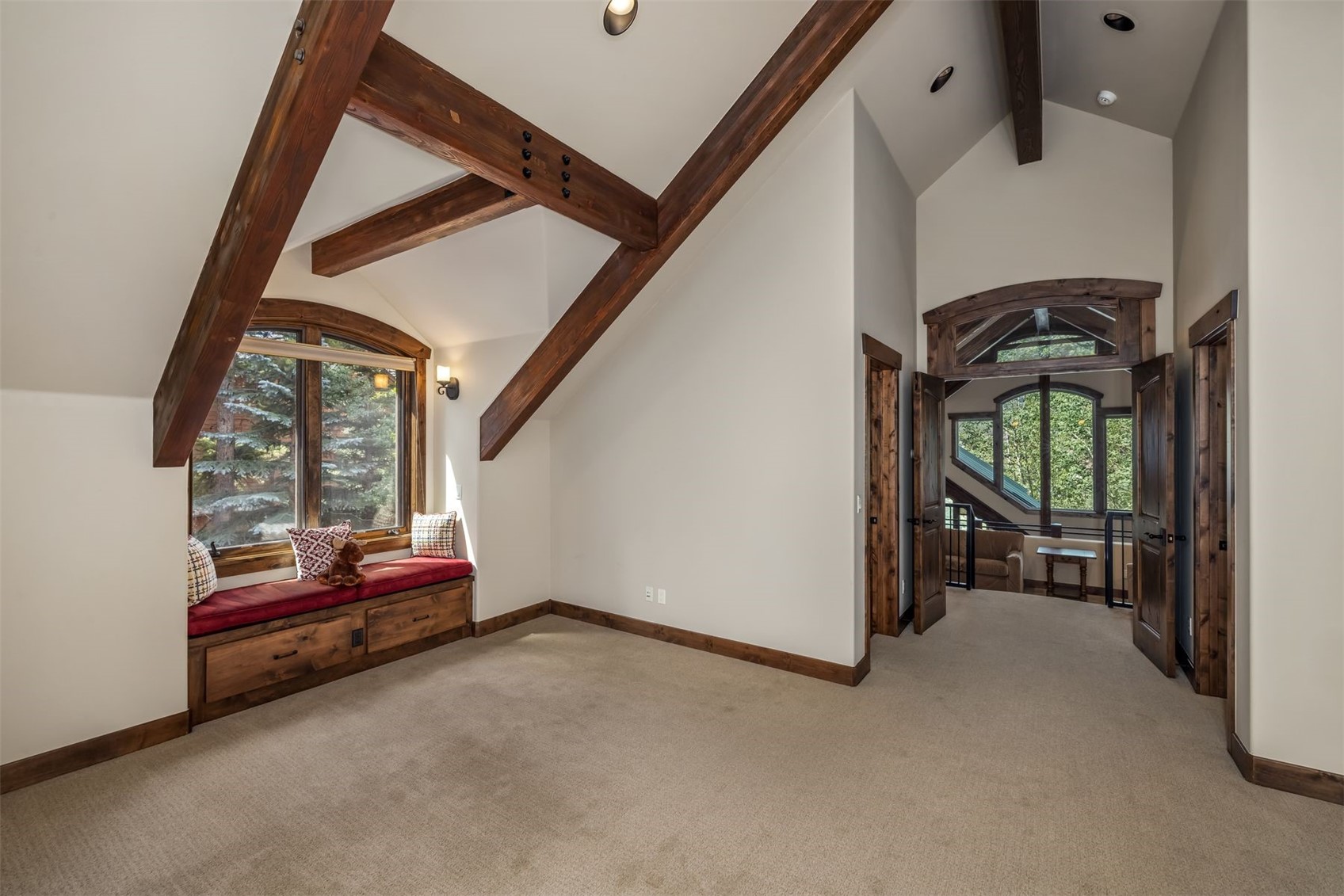 This cozy bedroom features a vaulted ceiling with exposed dark wood beams and a built-in window seat with storage drawers beneath. The room is carpeted and includes a doorway leading to an adjacent landing area with a large arched window. The warm wood trim and neutral wall tones create a rustic, inviting atmosphere.