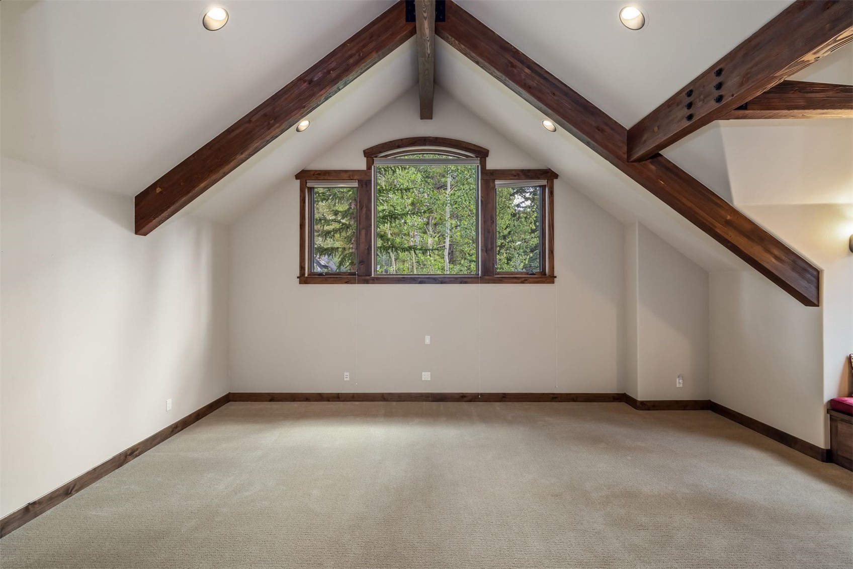 This spacious attic room features a vaulted ceiling accented by exposed dark wood beams, creating a rustic and cozy atmosphere. A central window assembly with a curved top provides a clear view of the surrounding trees, while recessed lighting illuminates the neutral-toned walls and carpeted floor. The room offers a versatile, open layout suitable for a bedroom, office, or recreational space.