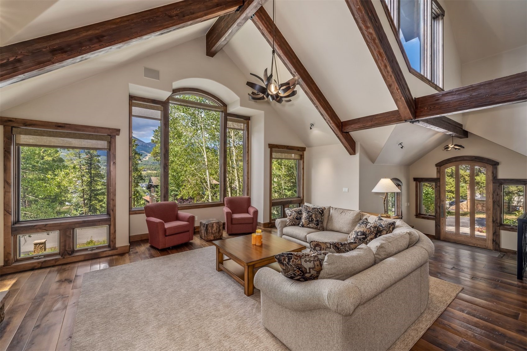 This spacious living room features a dramatic vaulted ceiling with exposed dark wood beams and a large, multi-pane window configuration that frames a scenic mountain view. The room is furnished with a neutral-toned sectional sofa and two red armchairs arranged around a central wooden coffee table on a large area rug. The warm, rustic aesthetic is complemented by dark wood flooring and a unique, dark-toned chandelier hanging from the peak of the ceiling.