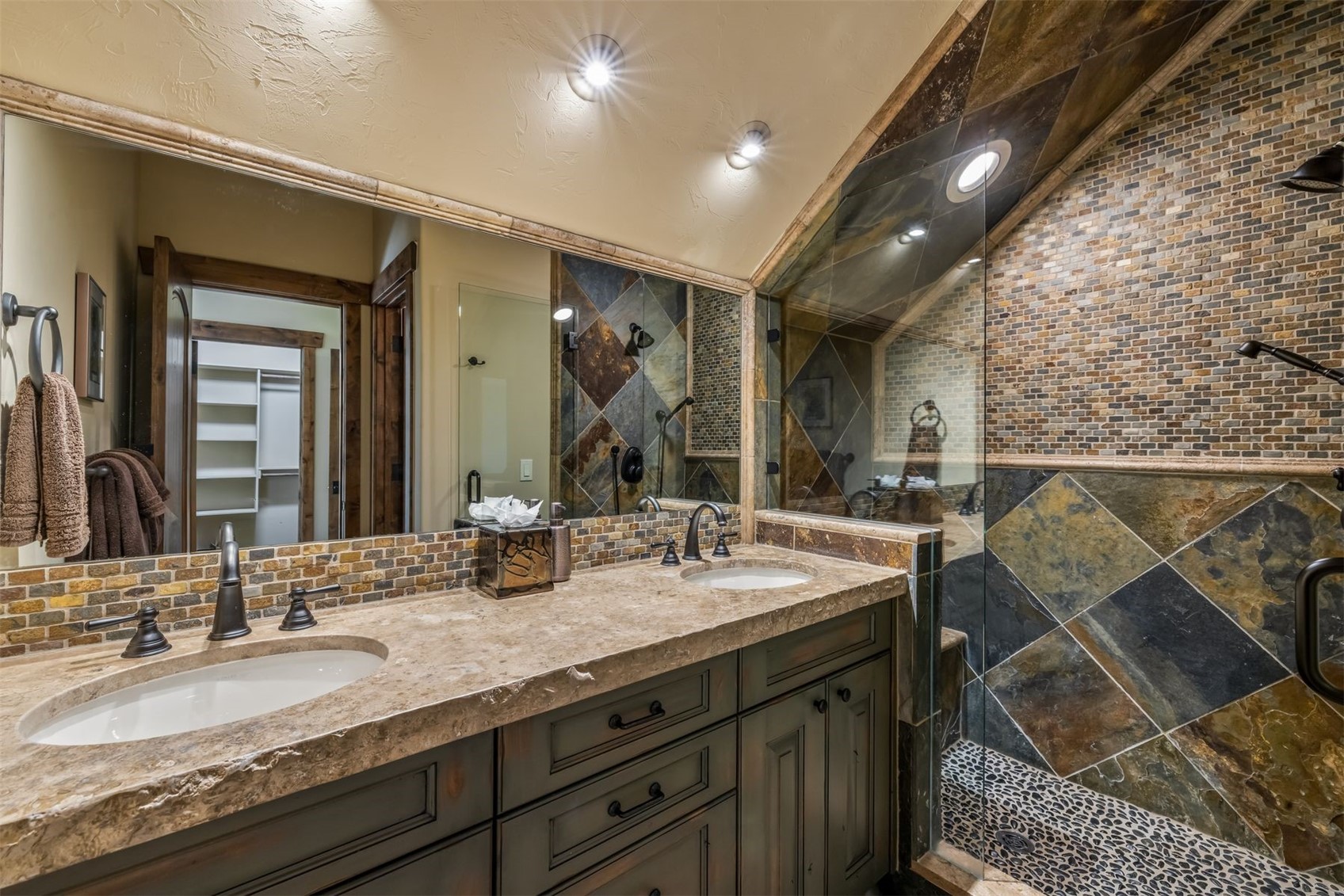 This primary bathroom features a dual-sink vanity with a stone countertop and dark cabinetry, complemented by a large mirror reflecting the space. To the right, a walk-in shower is enclosed by glass and showcases intricate stone tile work on the walls and a pebble-textured floor. The room is illuminated by recessed lighting, creating a warm and rustic atmosphere.