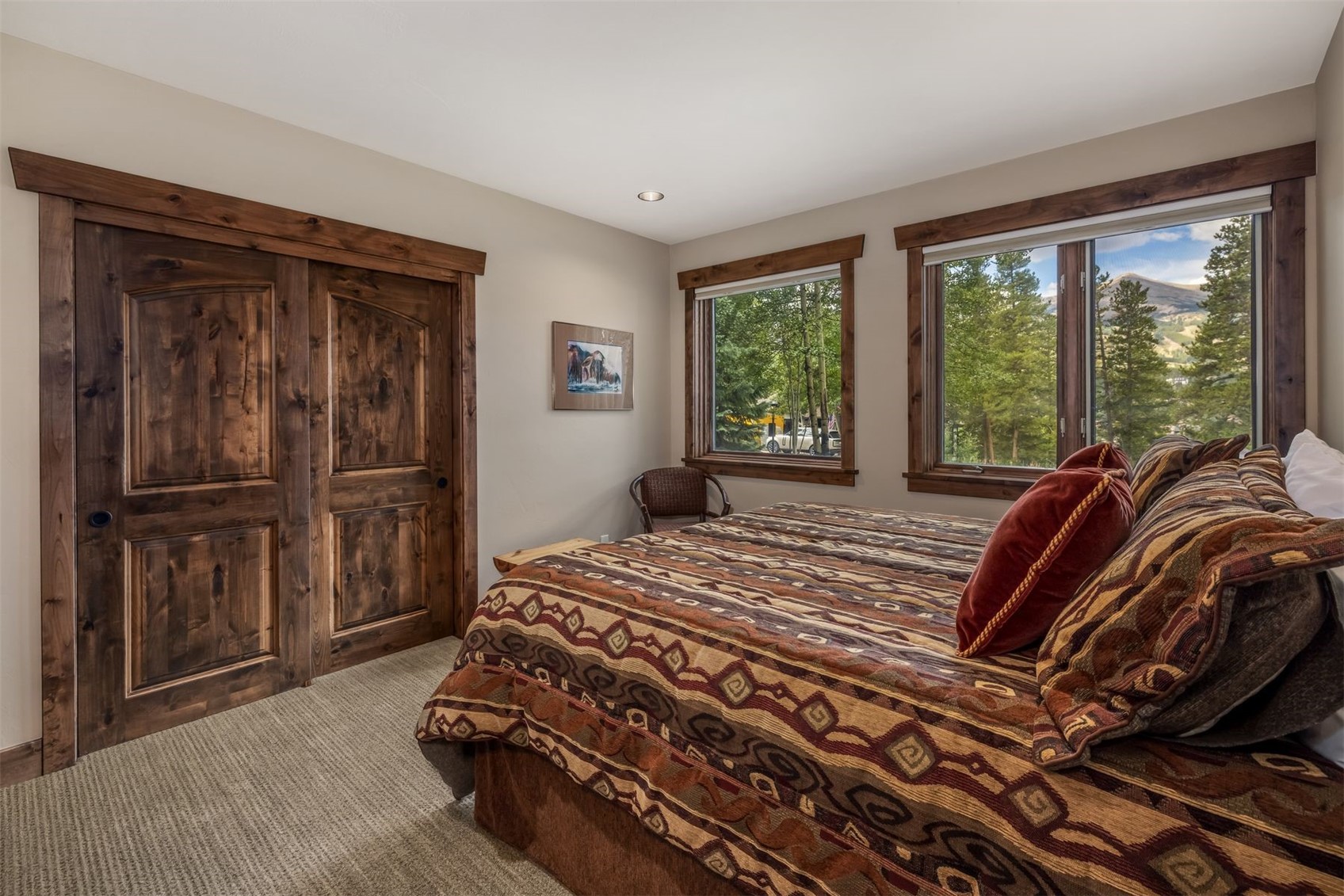 This cozy bedroom features a rustic aesthetic with dark wood trim around the doors and windows. A bed with a patterned, earth-toned quilt and plush pillows serves as the focal point, positioned near two windows that offer a scenic view of the surrounding trees and mountains. The room is carpeted and includes a small wooden chair in the corner, creating a warm and inviting atmosphere.