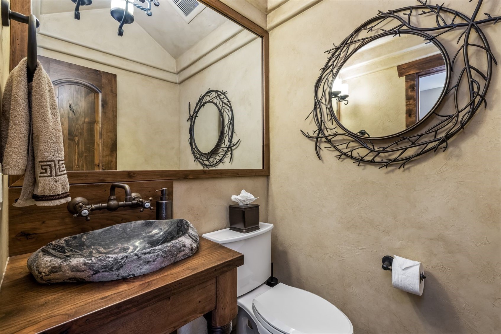 This rustic-style powder room features a unique stone vessel sink mounted on a natural wood vanity. The walls are finished in a textured, warm-toned plaster, complemented by a decorative twig-style mirror and a large rectangular mirror above the vanity. A white toilet and dark bronze fixtures complete the space, creating a cozy and earthy atmosphere.